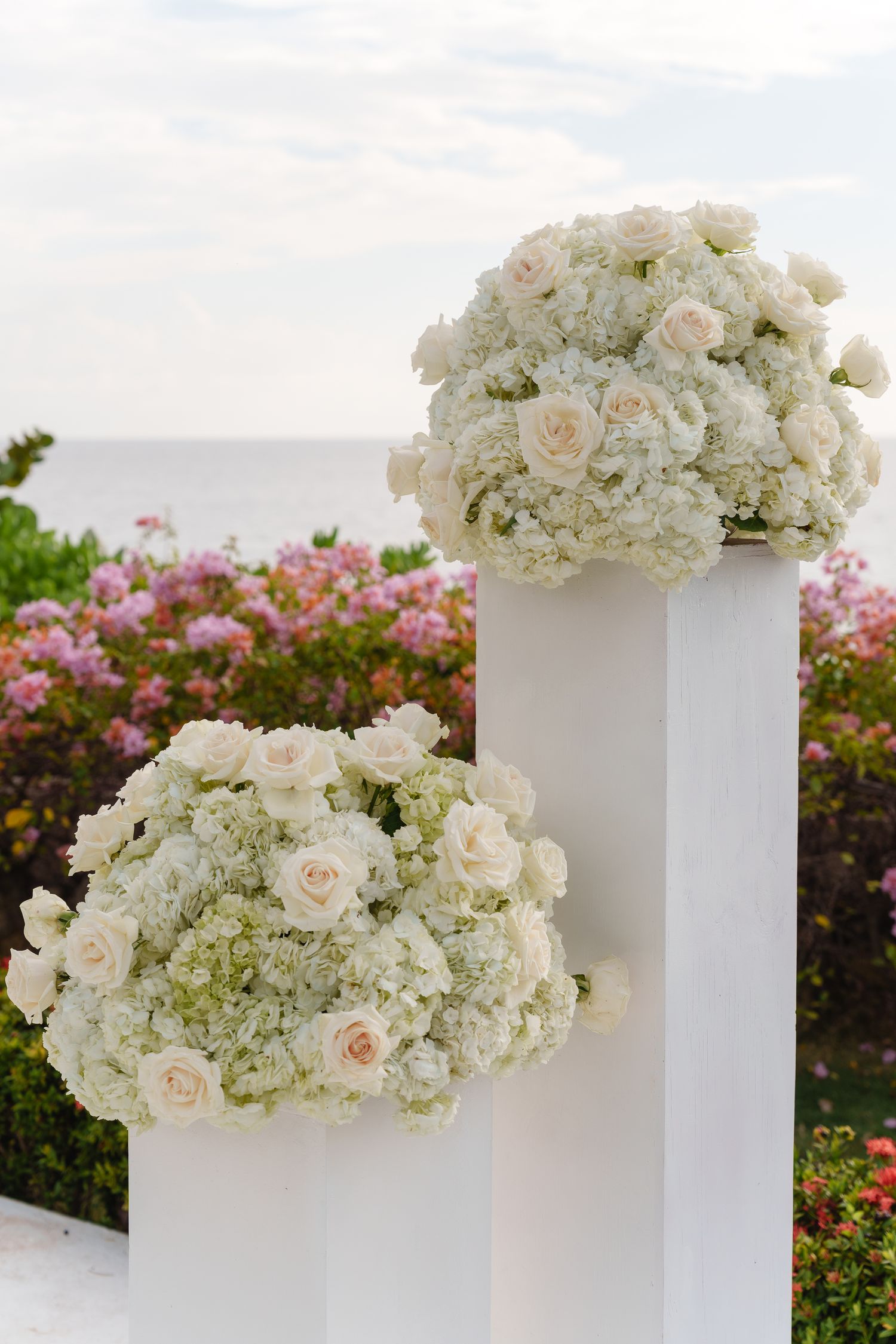 White floral arrangements with roses and hydrangeas displayed on tall pedestals against a scenic ocean backdrop with pink flowers.