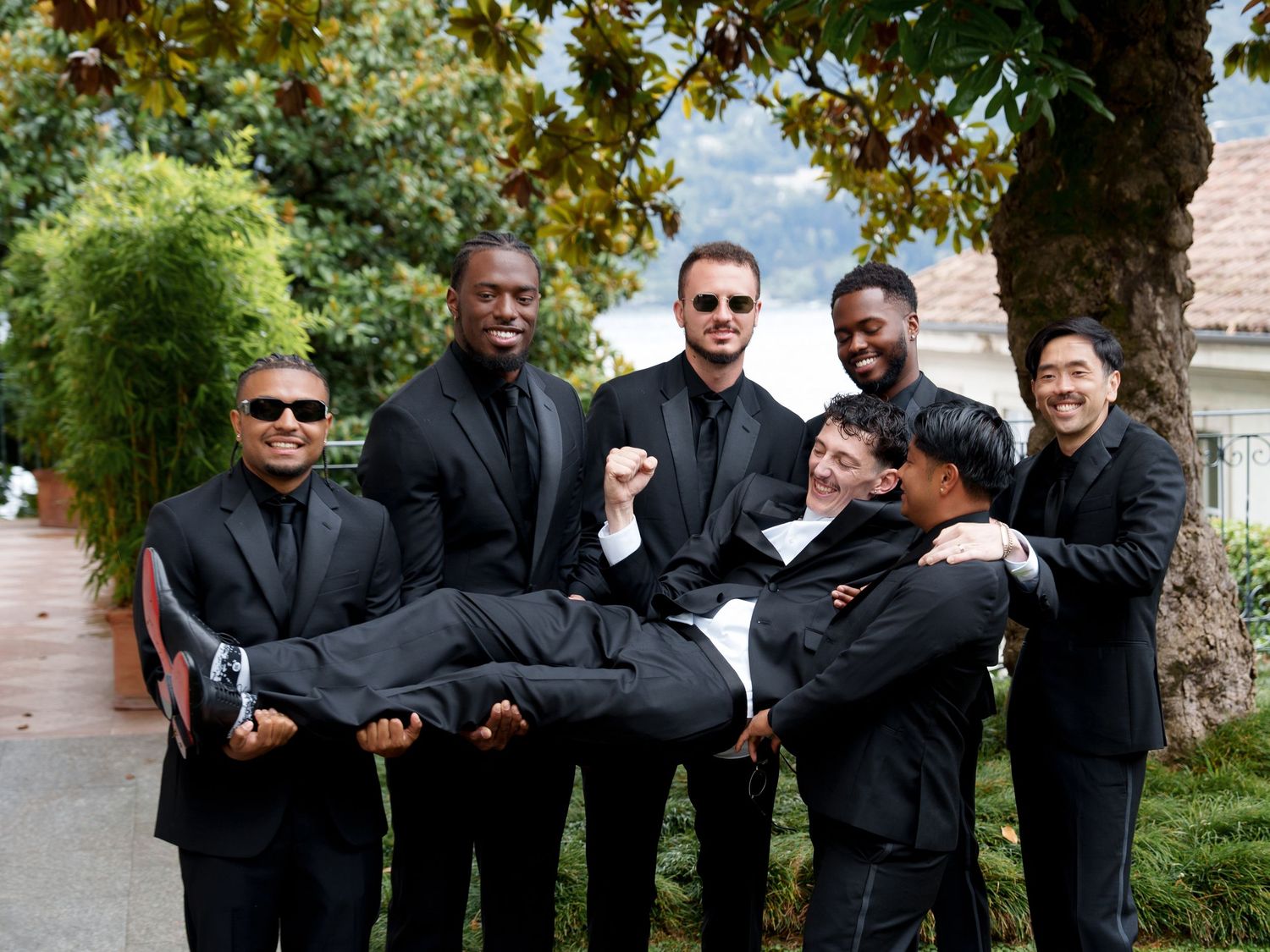 Groomsmen in black suits pose playfully for wedding photos outdoors on a sunny autumn day.