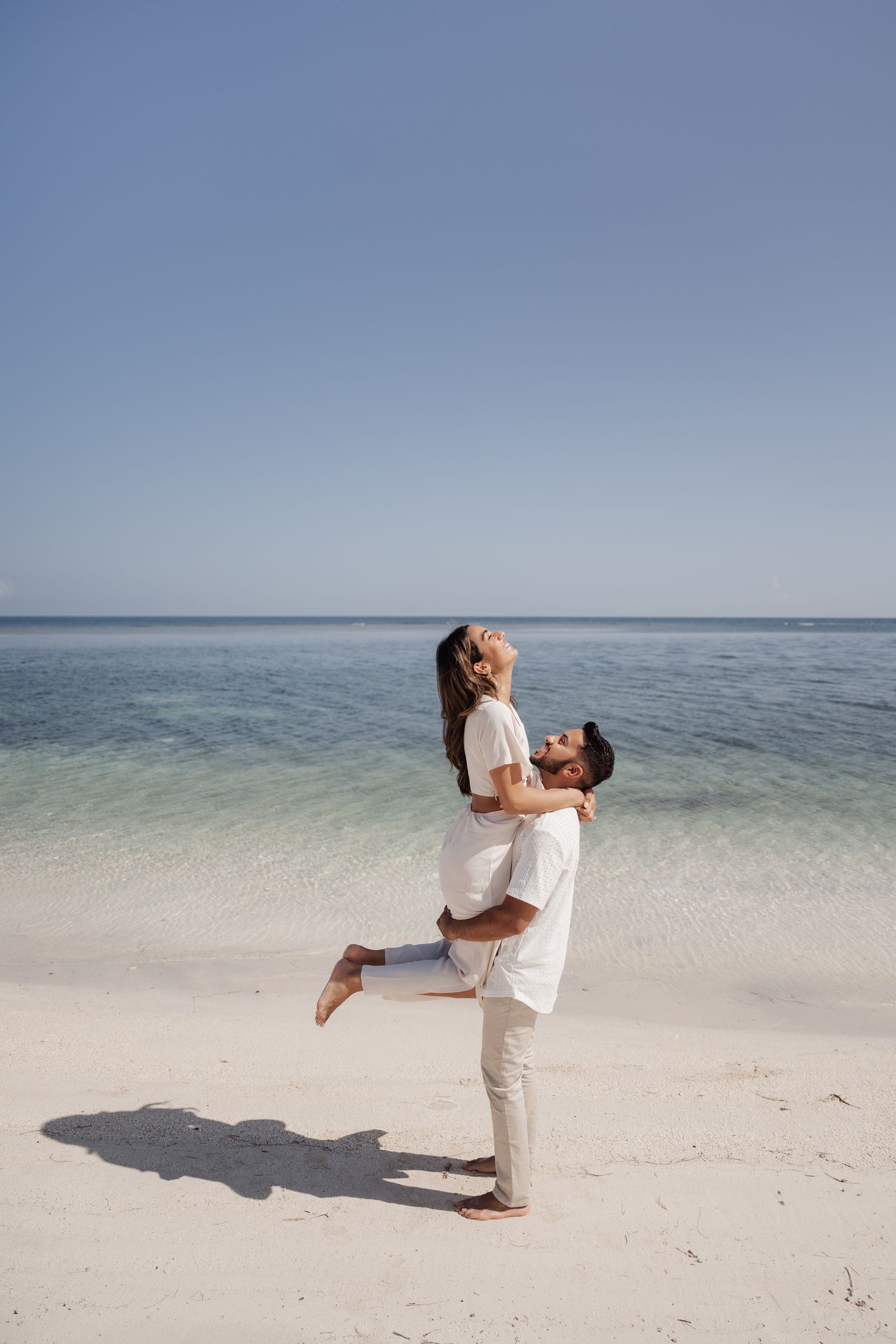 A romantic couple embraces on a pristine white sand beach with turquoise waters and clear blue sky in the background.