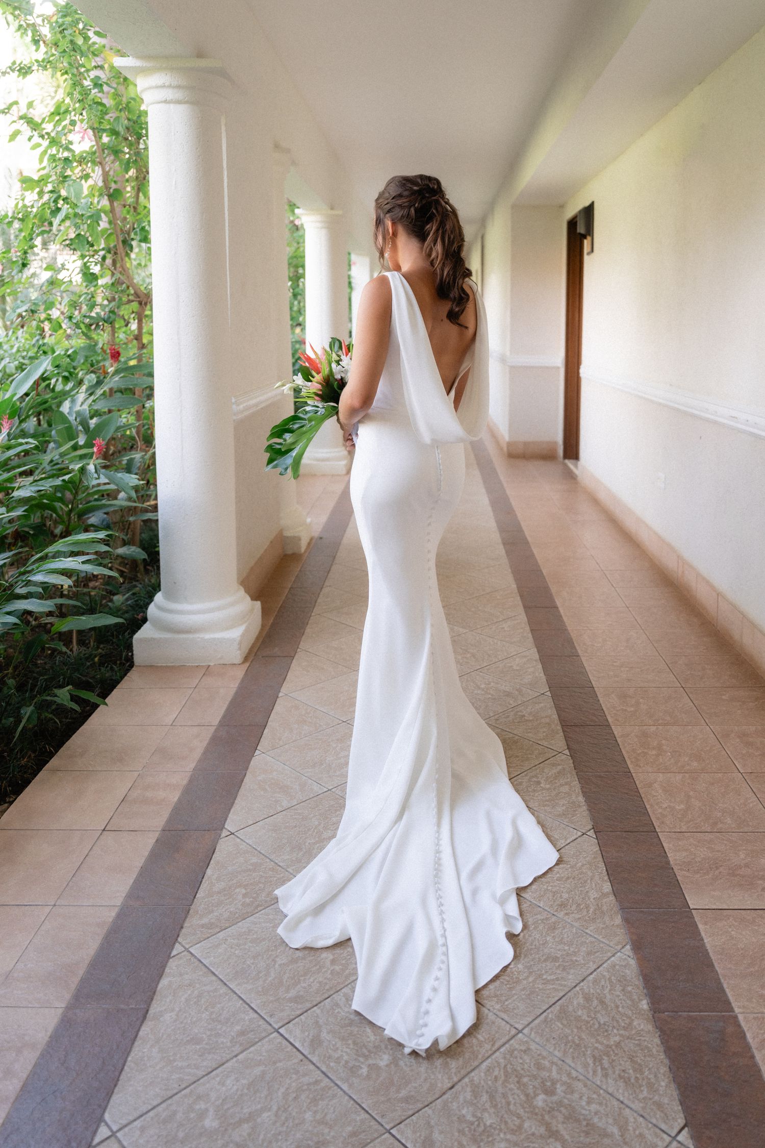Back view of bride in form-fitting white dress with train walking down columned hallway.