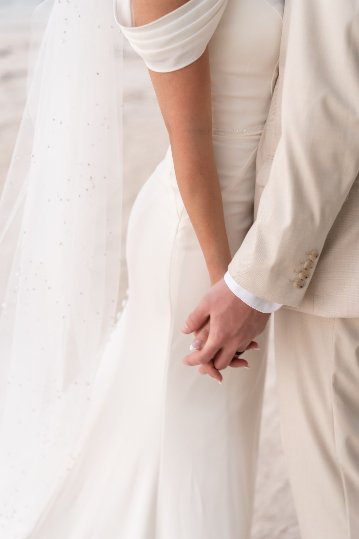 Close-up of newlyweds holding hands, showing white wedding dress and light colored suit details.