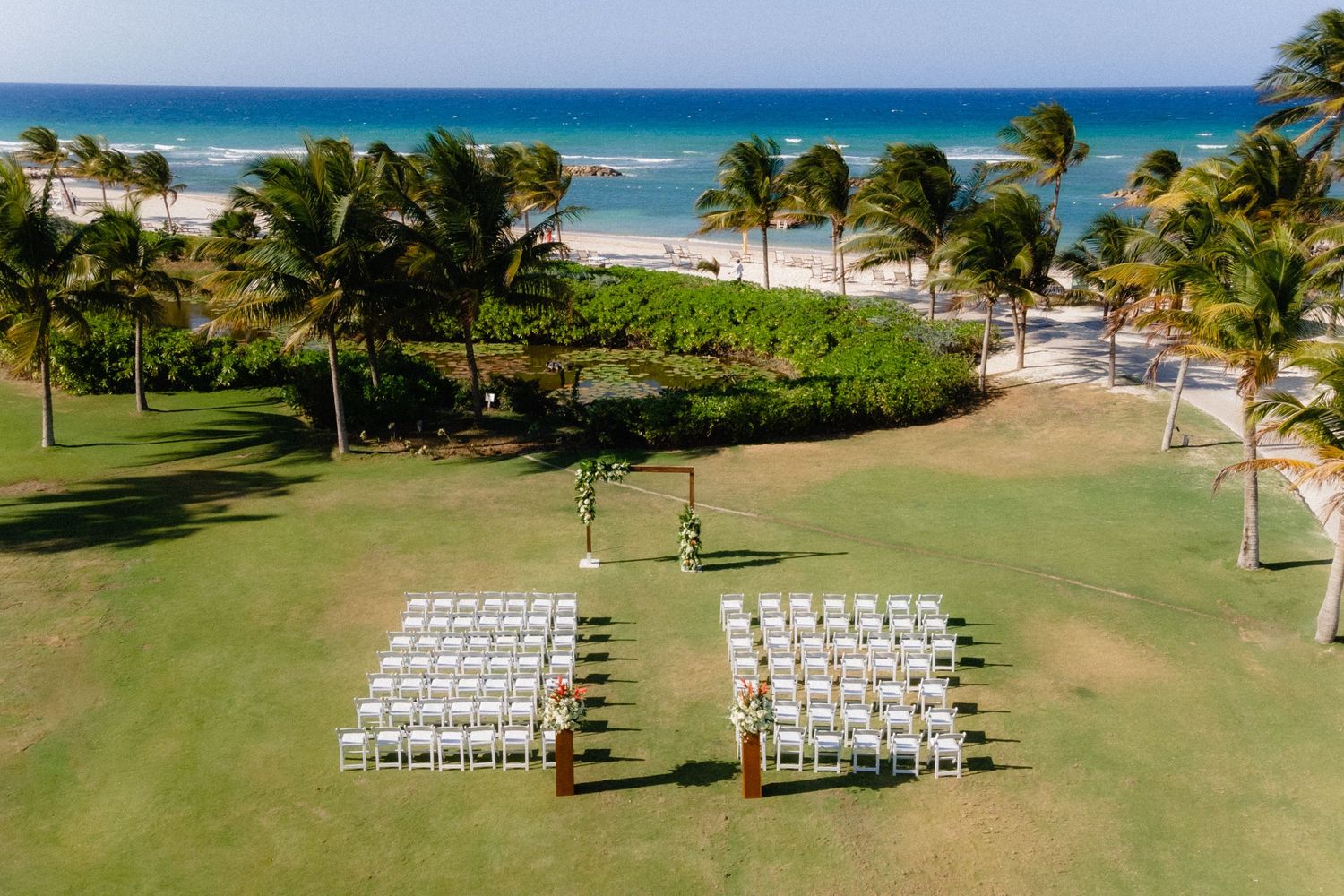 An aerial view of a beach wedding ceremony setup with white chairs on a tropical oceanfront lawn.