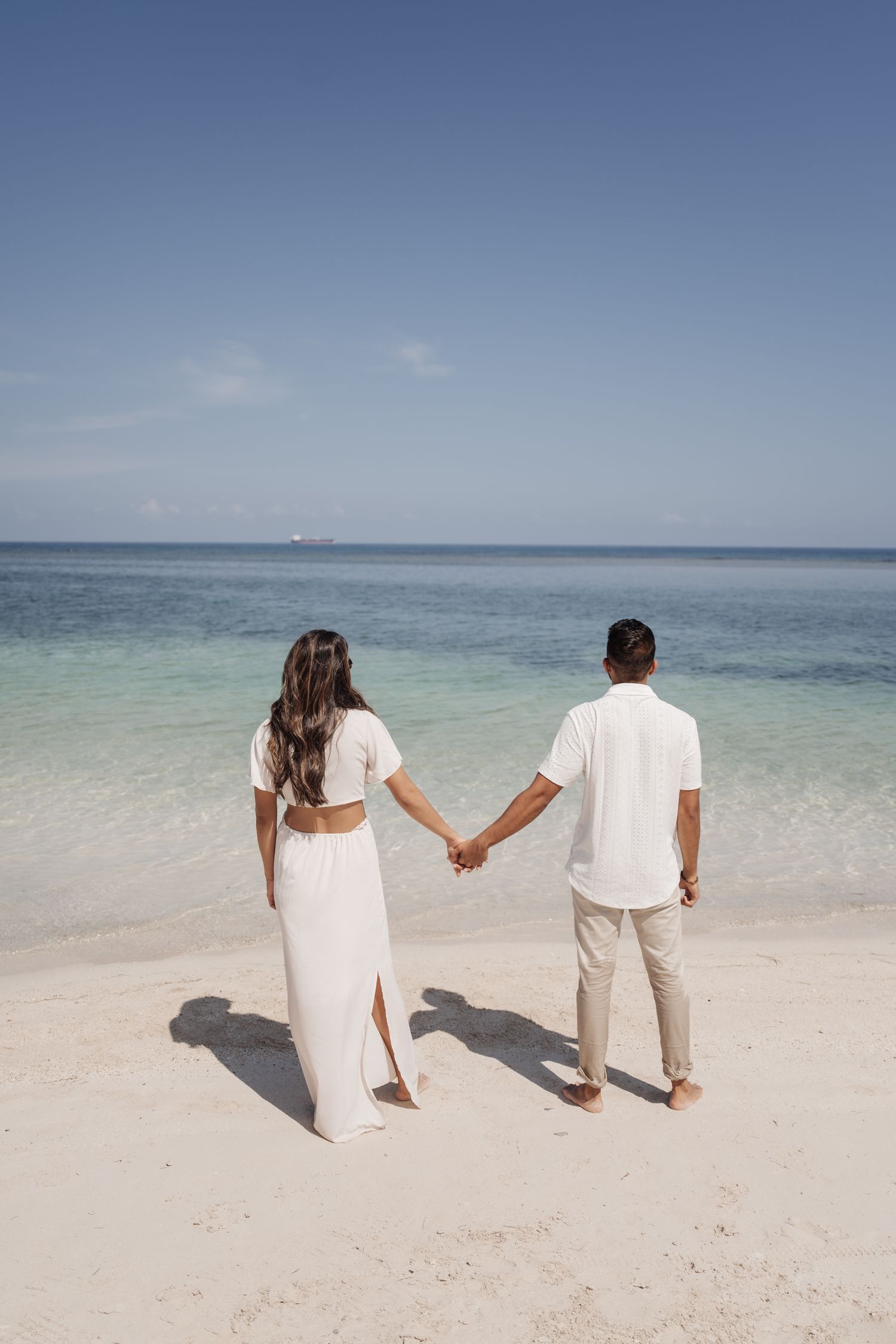 A couple holds hands while walking along a pristine beach shore with clear blue waters.