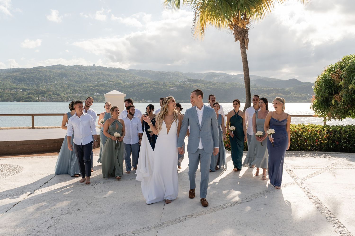 Wedding party walking together on a waterfront path with mountains in the background.