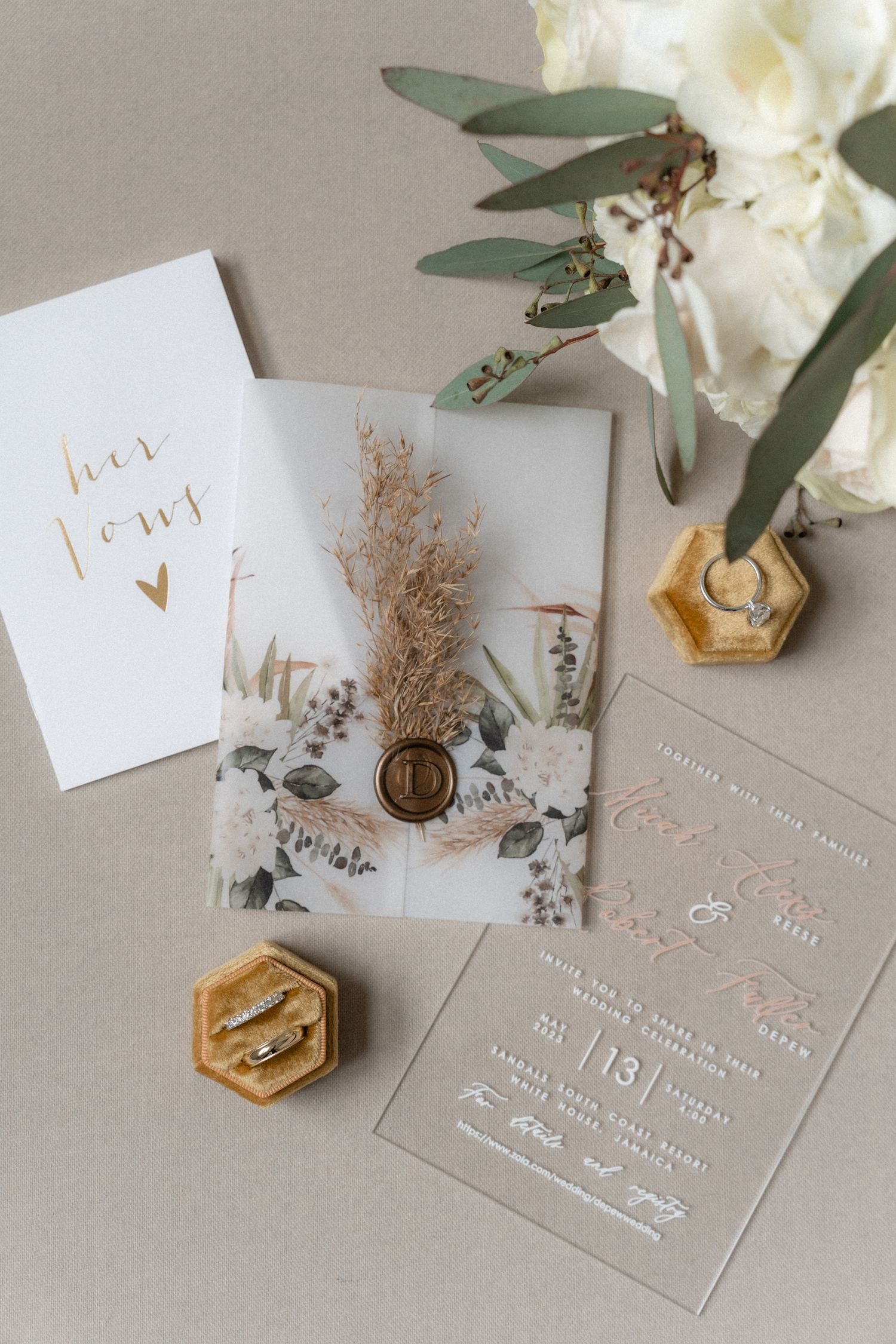 Elegant wedding invitation suite with gold lettering, dried flowers, and hexagonal wax seals on a neutral gray background.