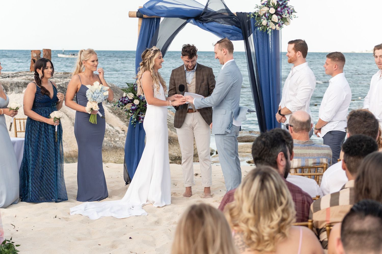 Oceanfront wedding ceremony with blue draped arch and attendants in coordinating attire.