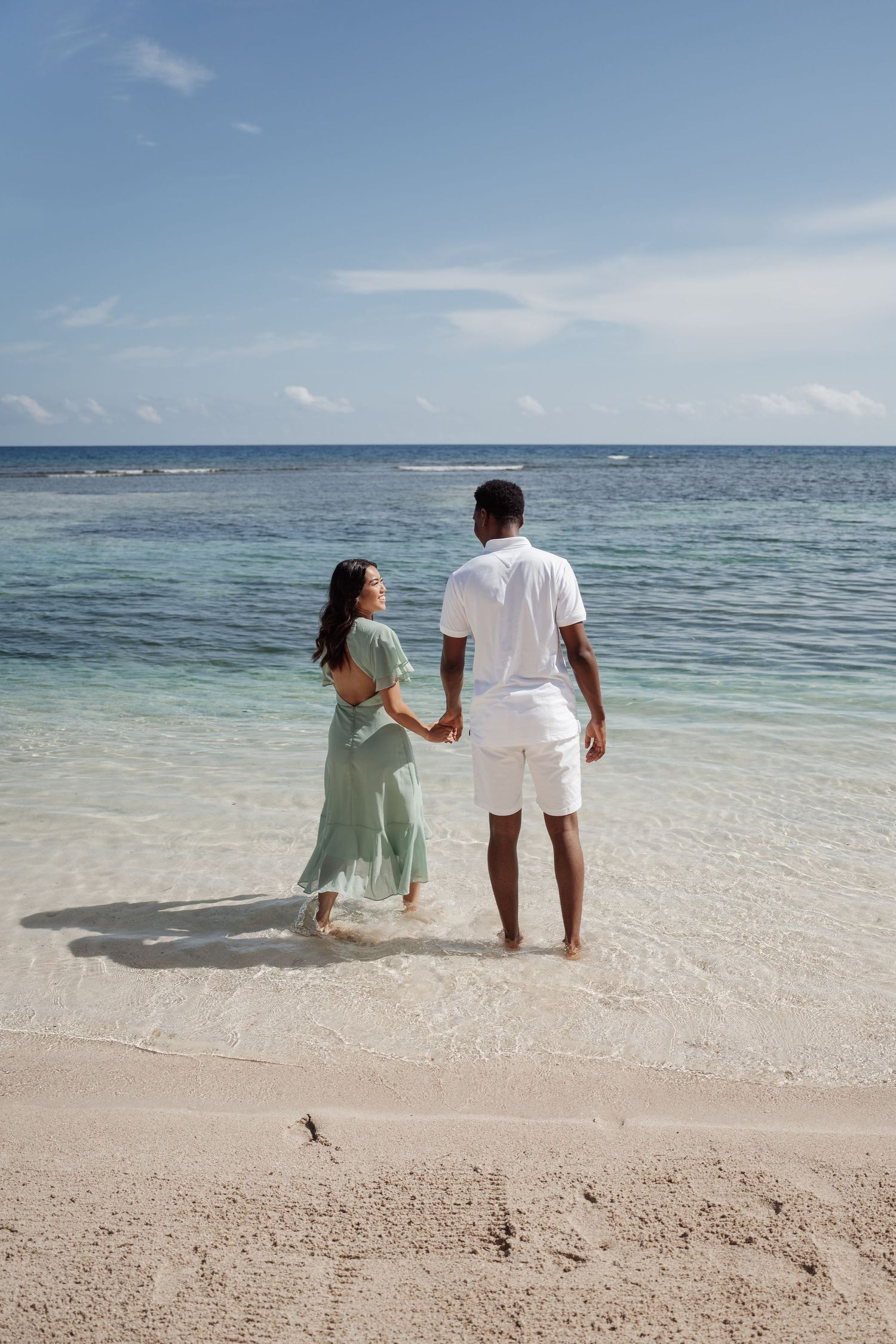 A couple holds hands while standing at the water's edge on a pristine beach.