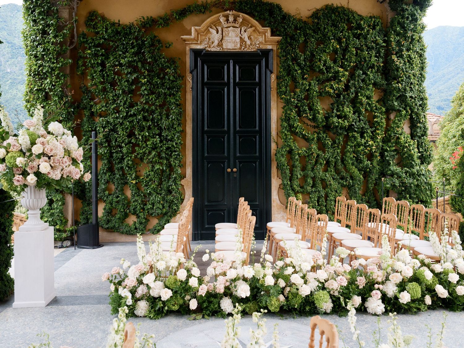 Elegant wedding ceremony setup with white floral arrangements against an ivy-covered wall with black doors.