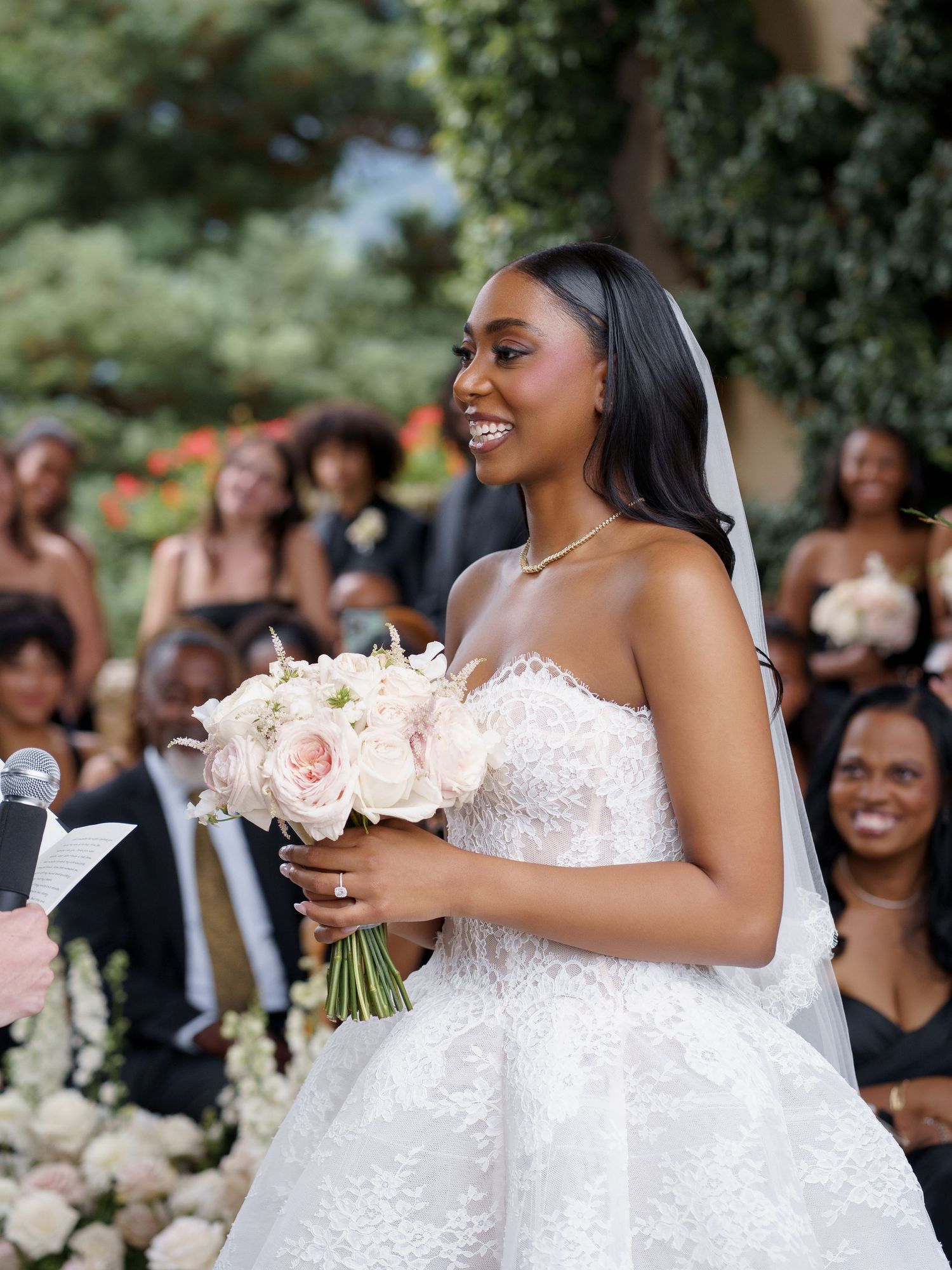 A bride in a white lace wedding gown holds a pink rose bouquet while smiling during an outdoor ceremony.