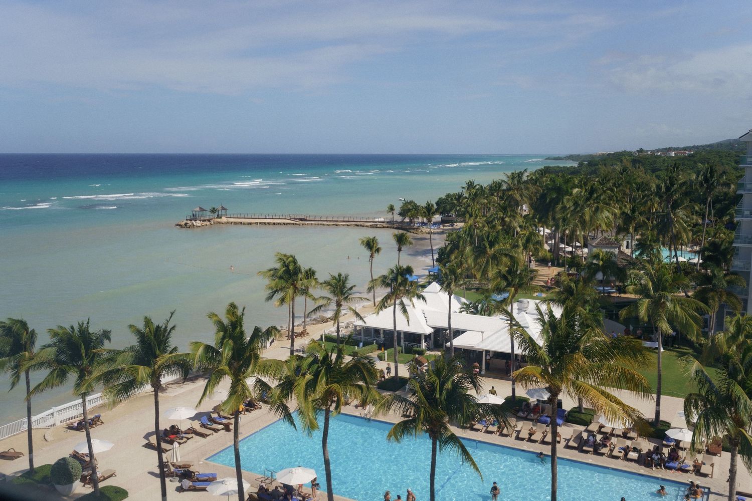 Tropical resort aerial view showing beachfront pool, palm trees and turquoise ocean waters.