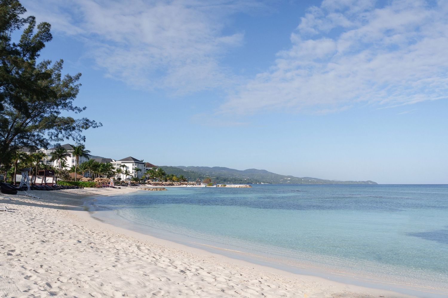 A serene white sand beach with clear turquoise waters and mountains in the distance in Jamaica.
