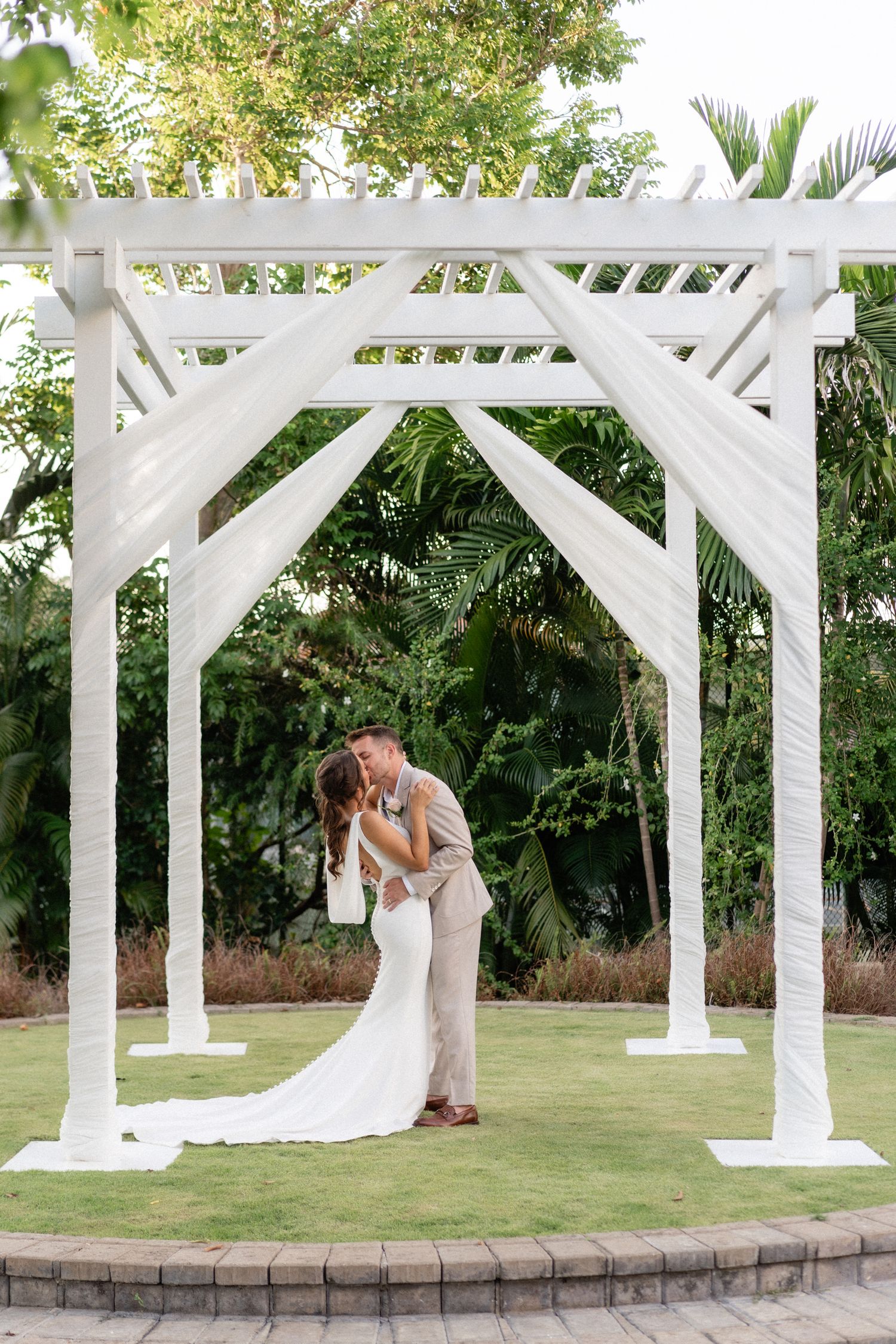 Newlyweds share romantic moment under white wooden pergola during garden wedding ceremony.