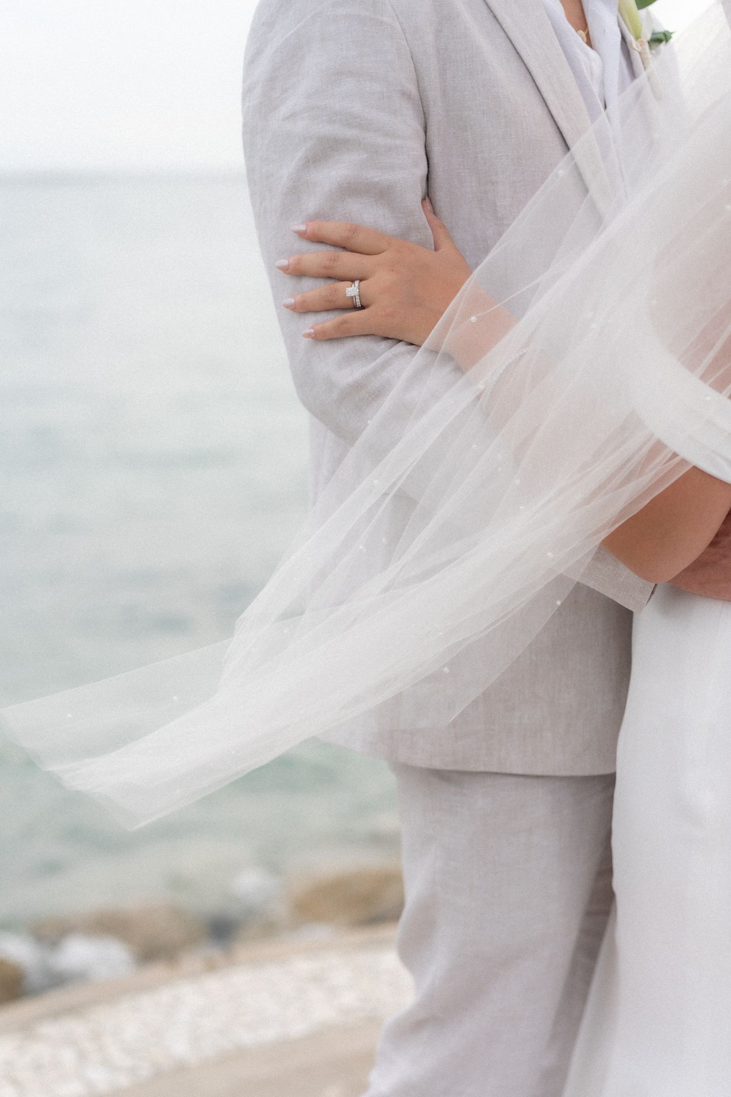 Close-up of wedding attire showing light gray suit and flowing white veil against ocean backdrop.
