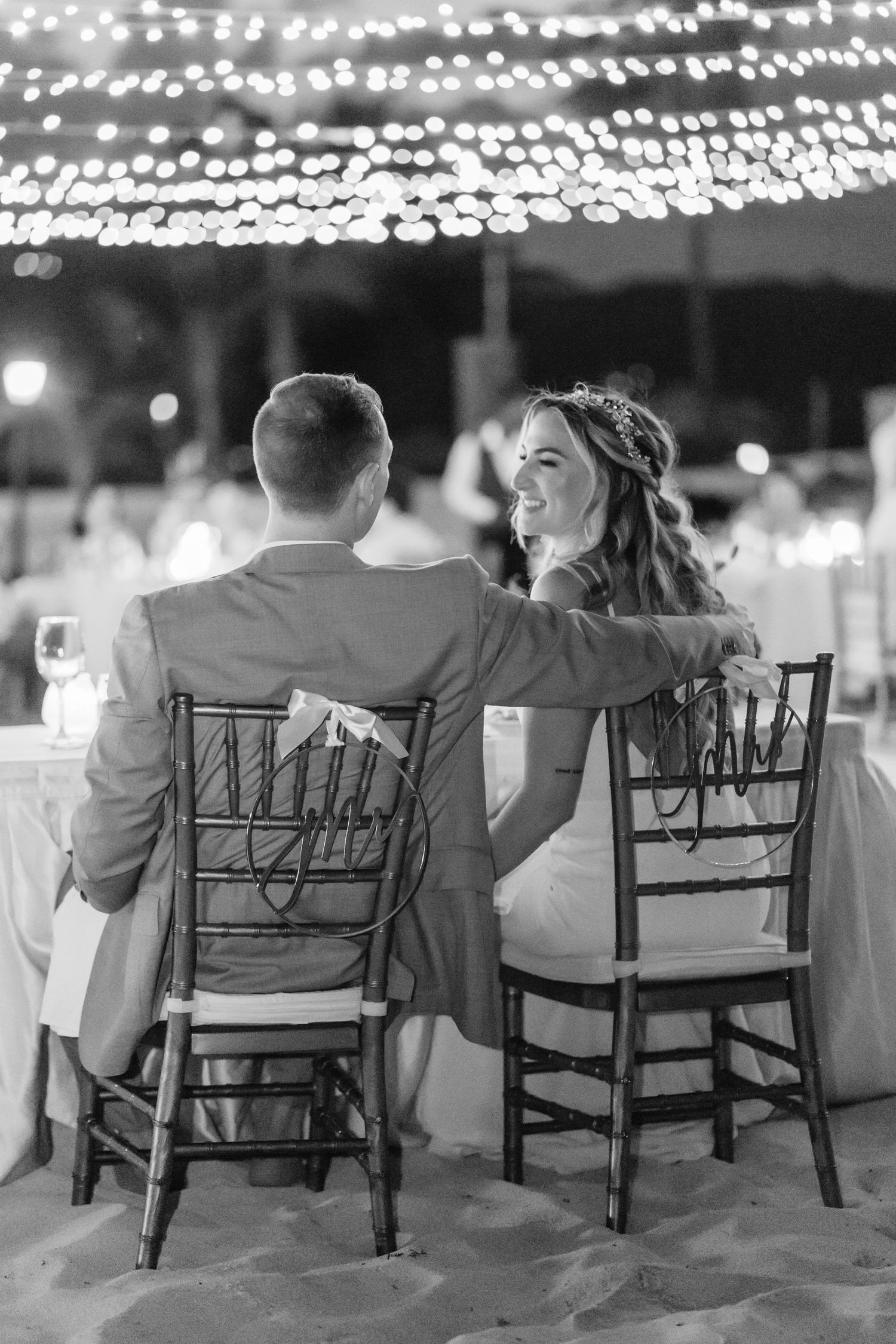 Black and white photo of couple at reception under twinkling lights sitting in Chiavari chairs.