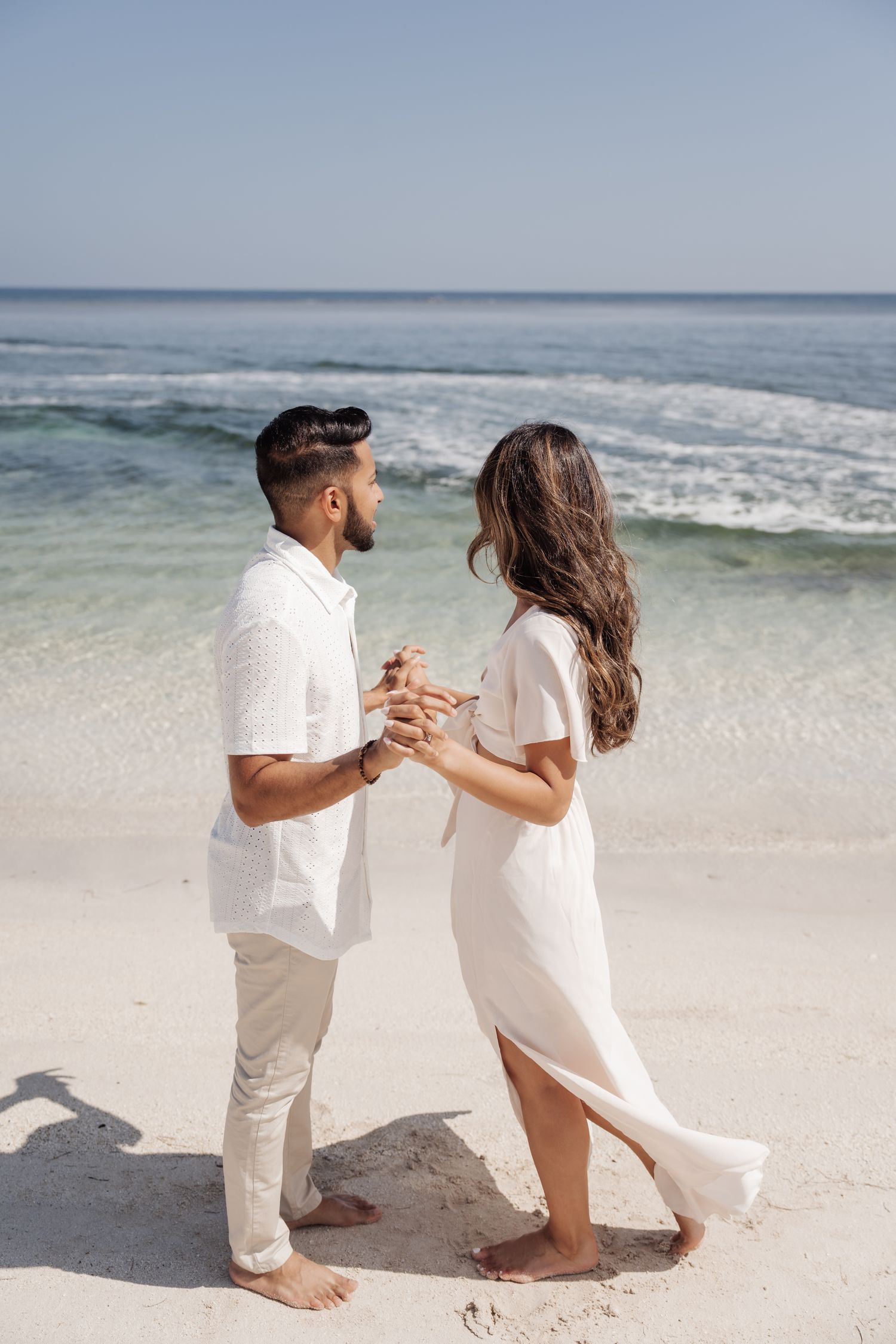 Couple in matching white outfits hold hands on sandy beach.
