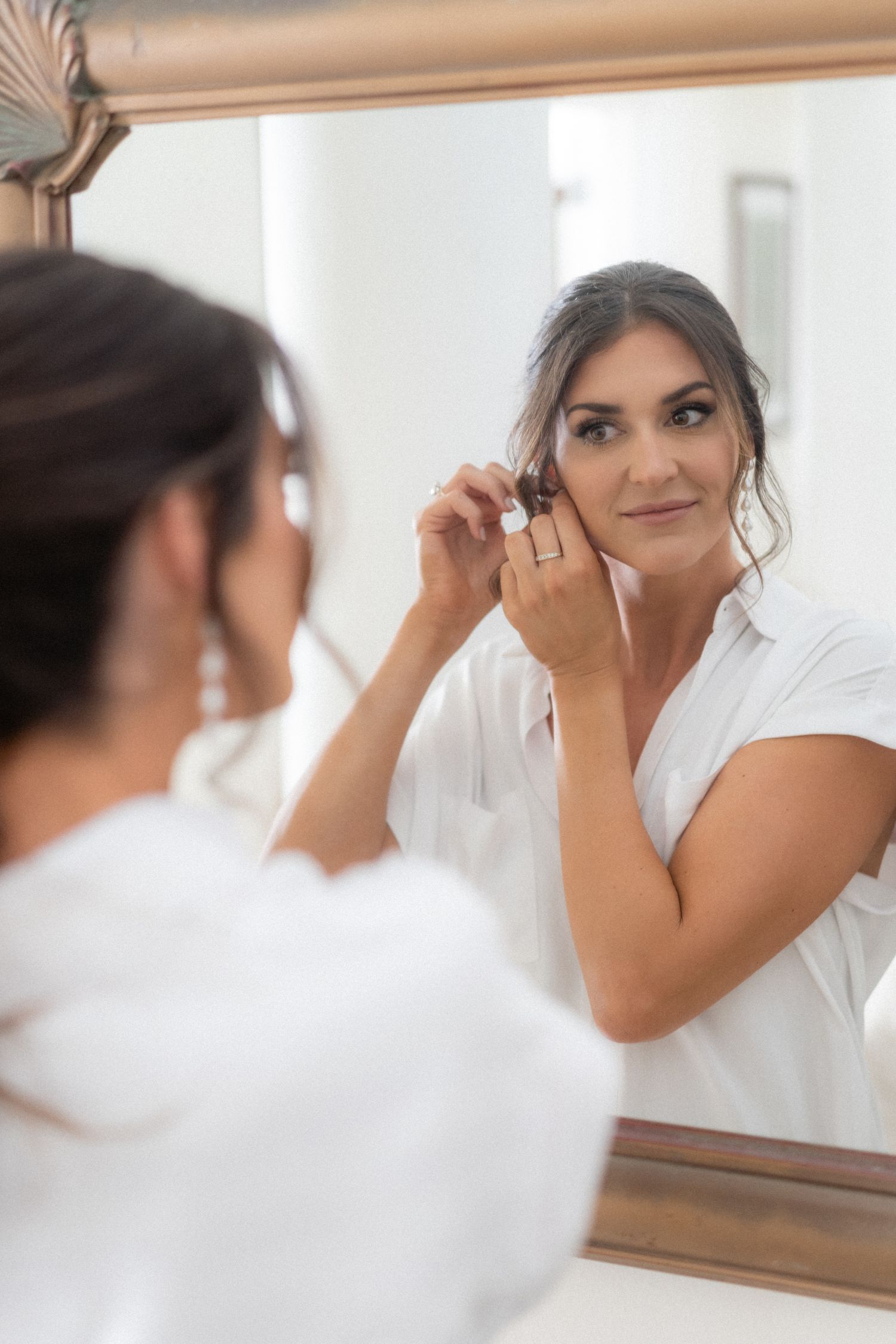 Getting ready scene of putting on earrings while looking in a mirror wearing a white robe.
