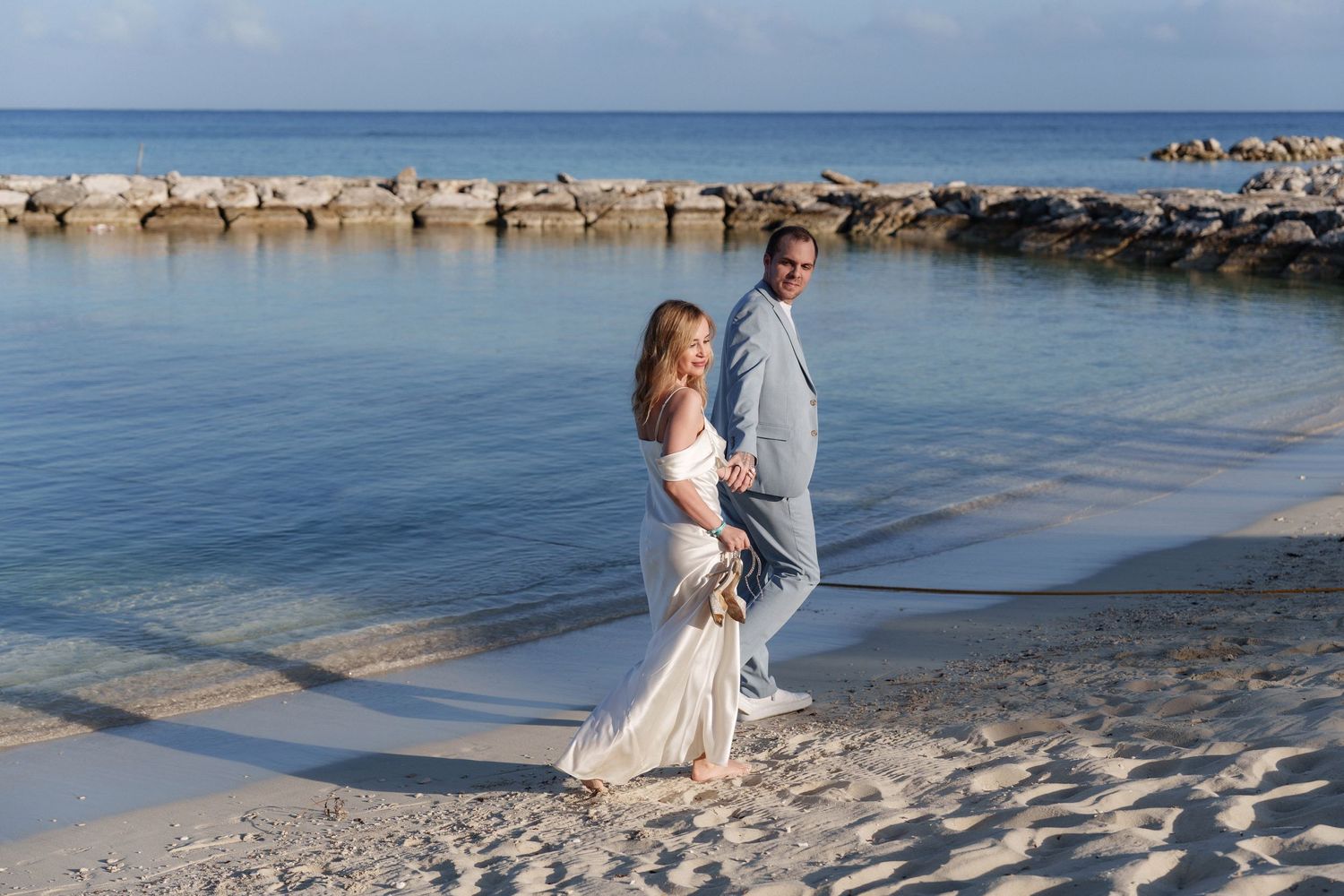 A couple in formal attire walking along a pristine beach with a stone jetty in background.
