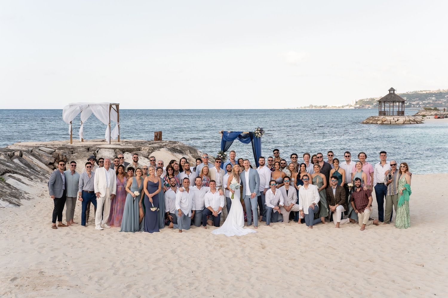 Large group beach wedding photo with guests gathered on sandy shore by the ocean.