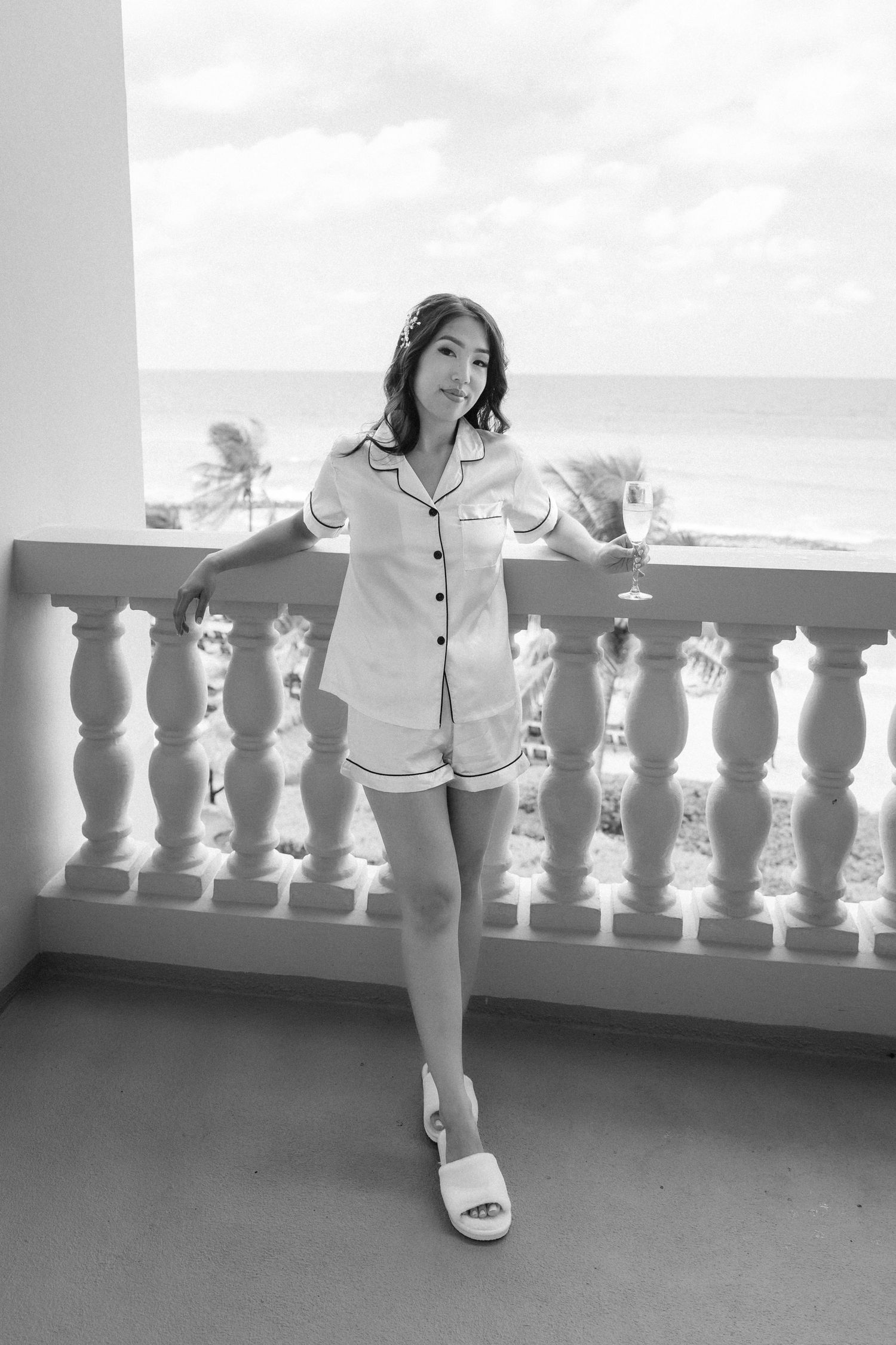 Black and white photo of someone in pajamas posing on a balcony overlooking the ocean.