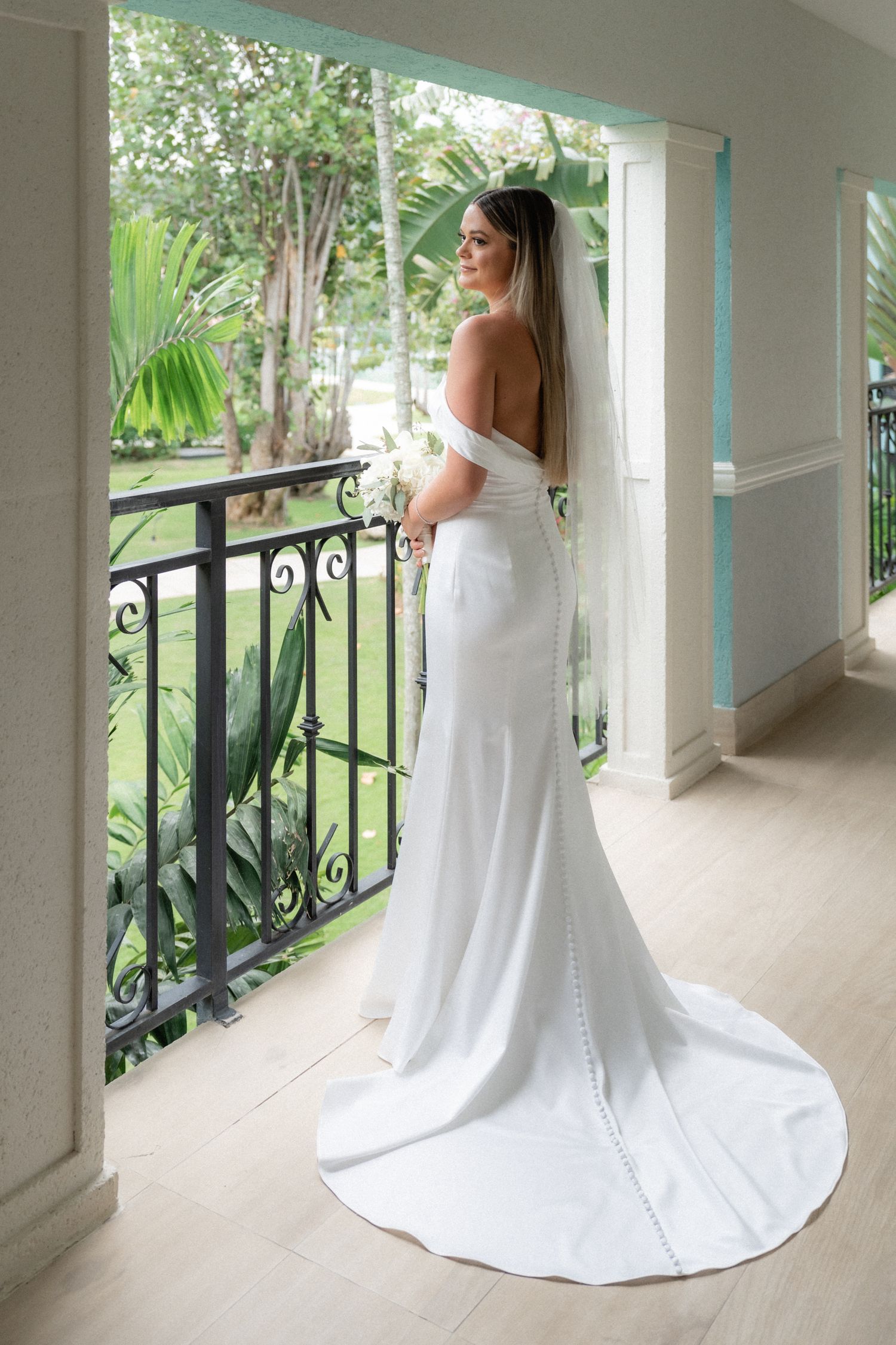 Elegant white wedding dress with a dramatic train photographed from behind on a tropical balcony.