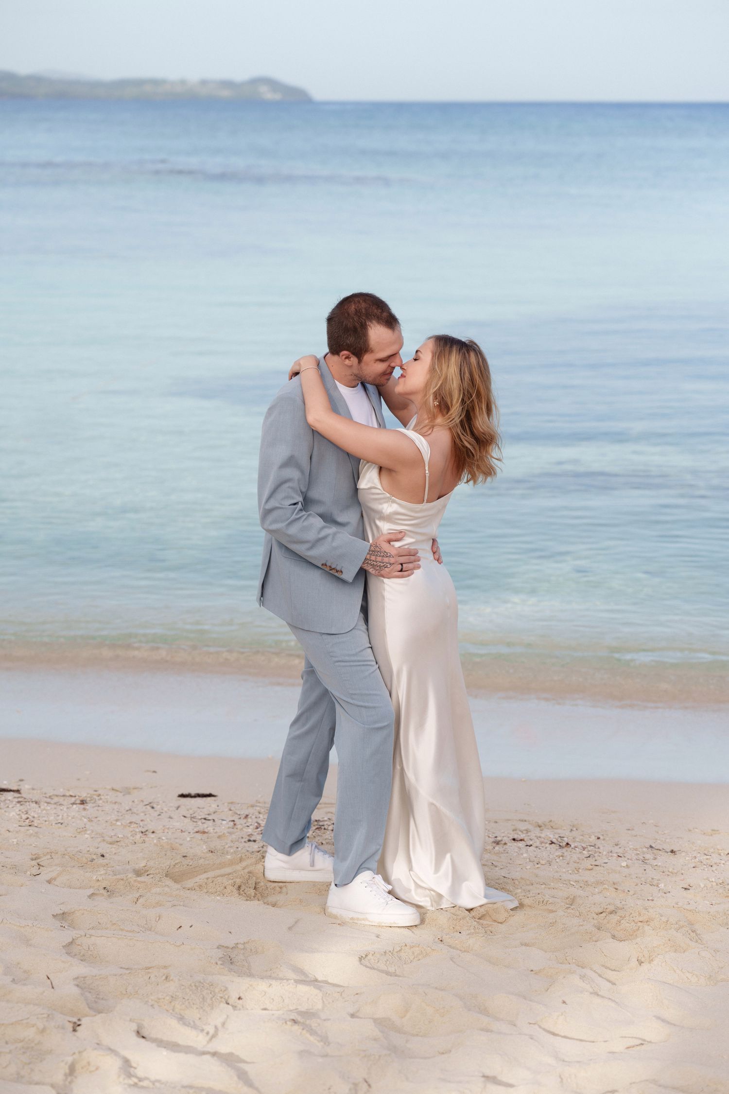 Couple embracing on sandy beach in formal wear with ocean backdrop.