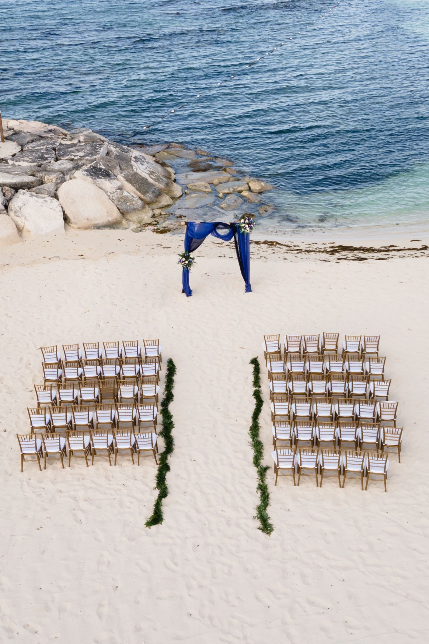 Aerial view of beach wedding setup with wooden chairs arranged in rows and blue arch on sandy shore.