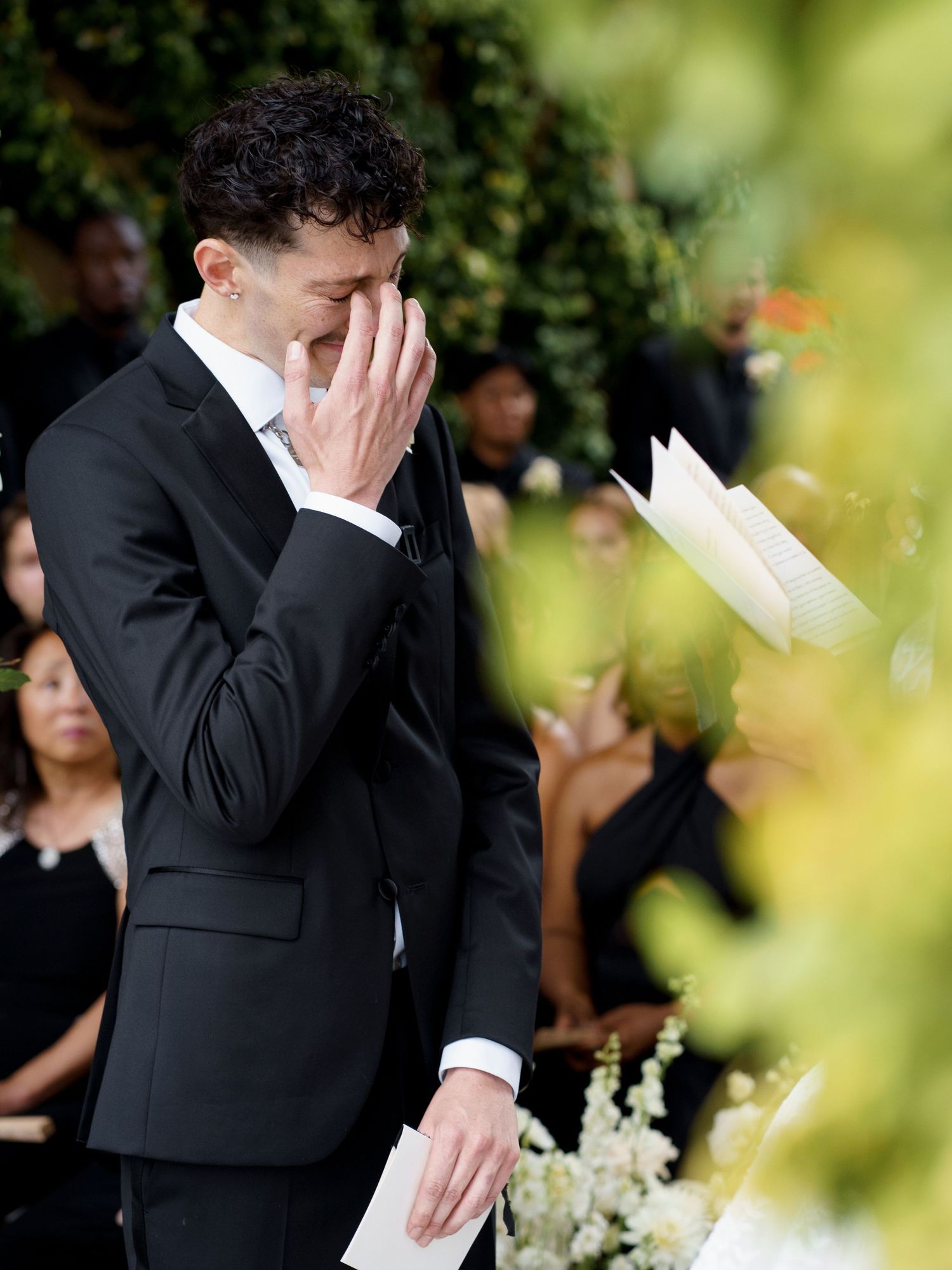 A person in a black suit dabs their eyes with a tissue during an emotional outdoor ceremony.