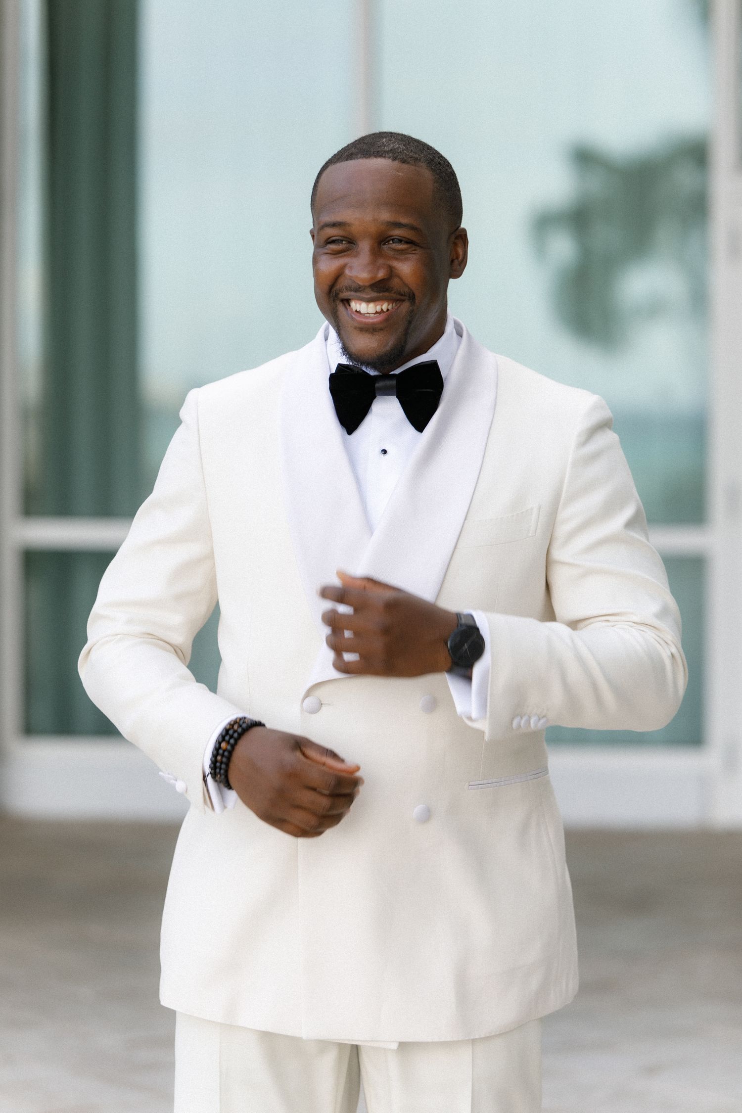 Groom in crisp white tuxedo smiles against modern architectural background.