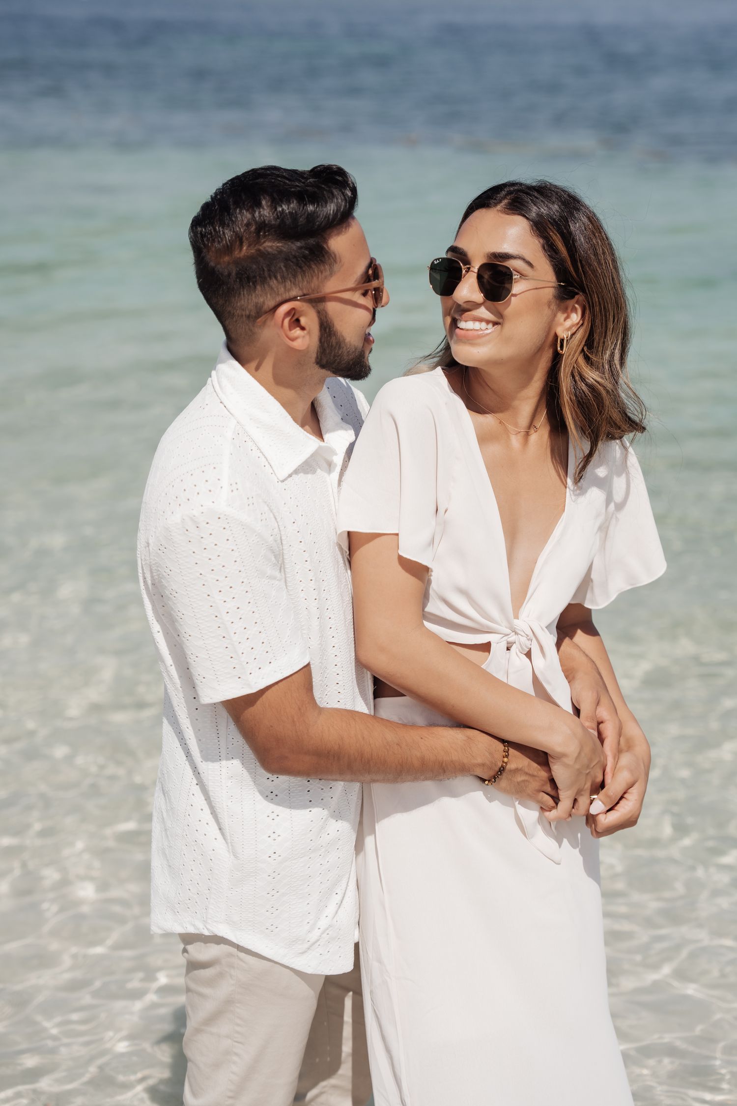 A couple in white summer clothing embracing on a beautiful beach with turquoise water.