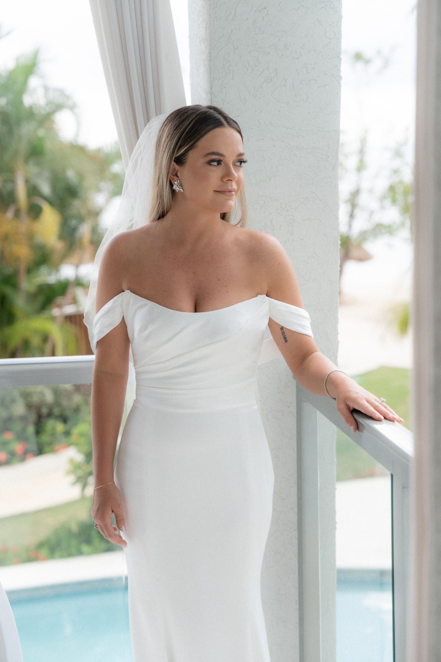 Elegant off-shoulder wedding dress detail with pool and tropical scenery in background.