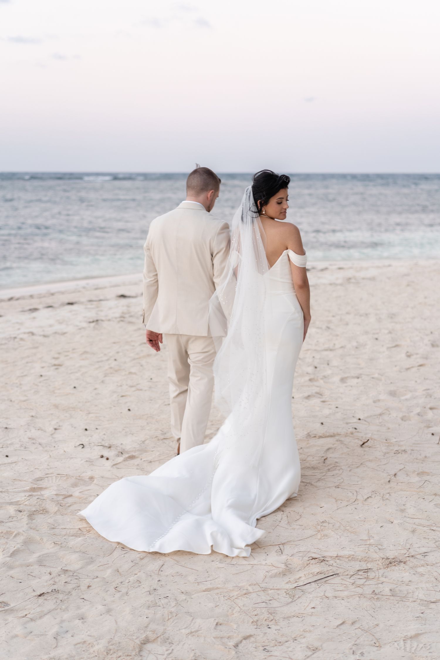 A wedding couple walks along a pristine beach at sunset in elegant white and beige attire.