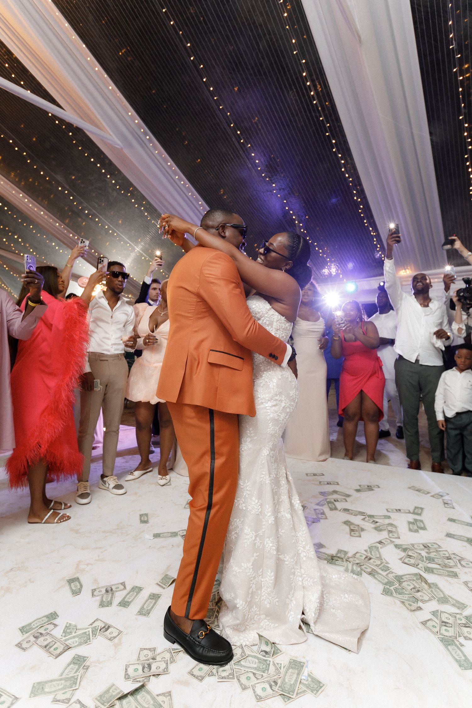 Wedding dance celebration with money scattered on floor under illuminated tent ceiling.