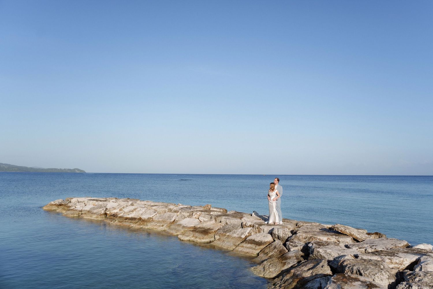 A figure in white stands on a rocky jetty overlooking calm blue waters.
