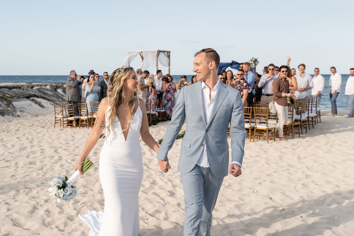 Newlyweds walk hand in hand on beach with wedding guests and blue ocean in background.