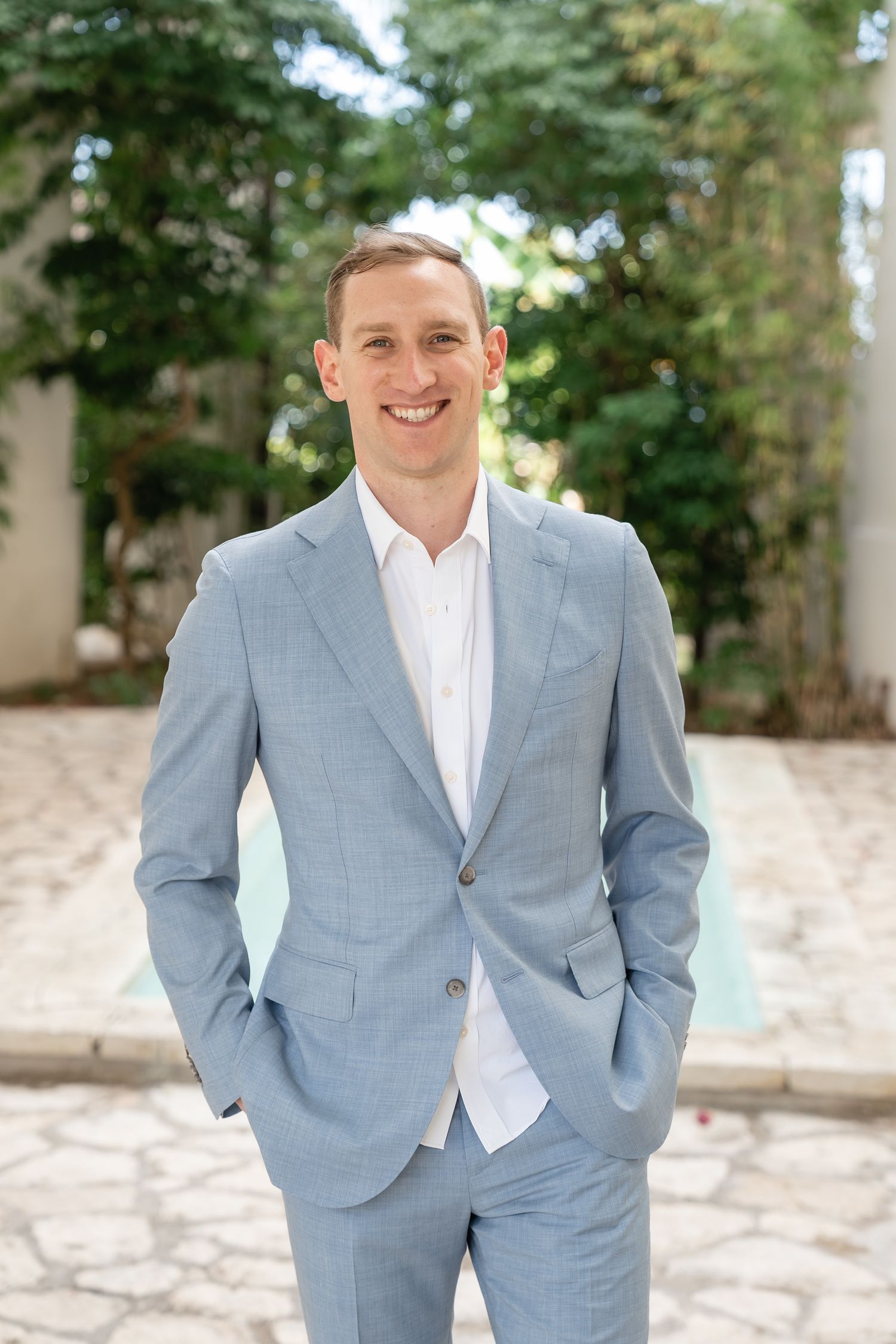 A person in a light blue suit and white dress shirt stands outdoors with a relaxed pose.