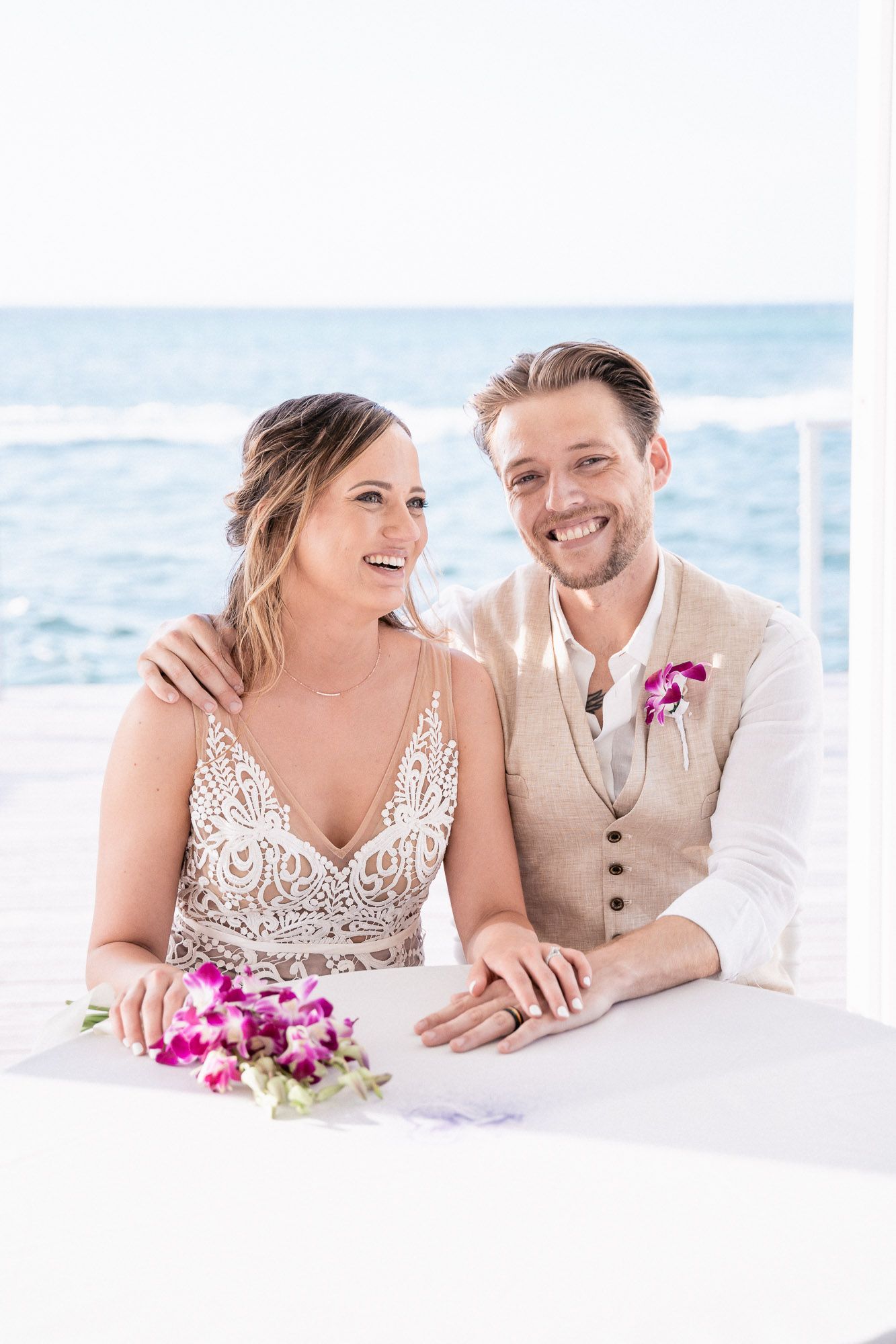 A joyful wedding portrait with bright ocean backdrop and pink floral accents.