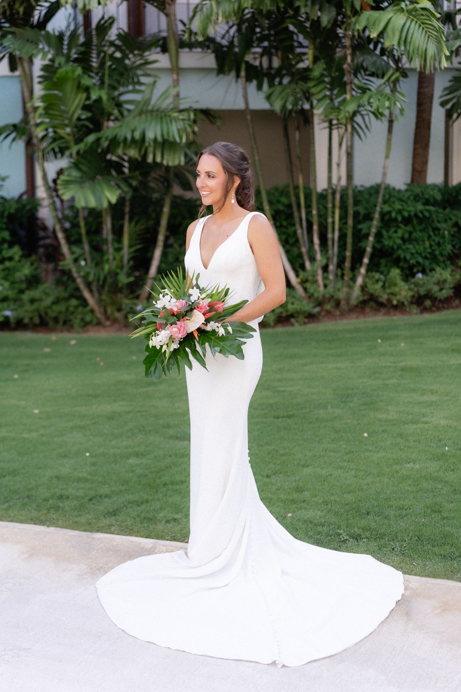 Elegant bridal portrait in a sleek white gown with tropical bouquet on manicured lawn.