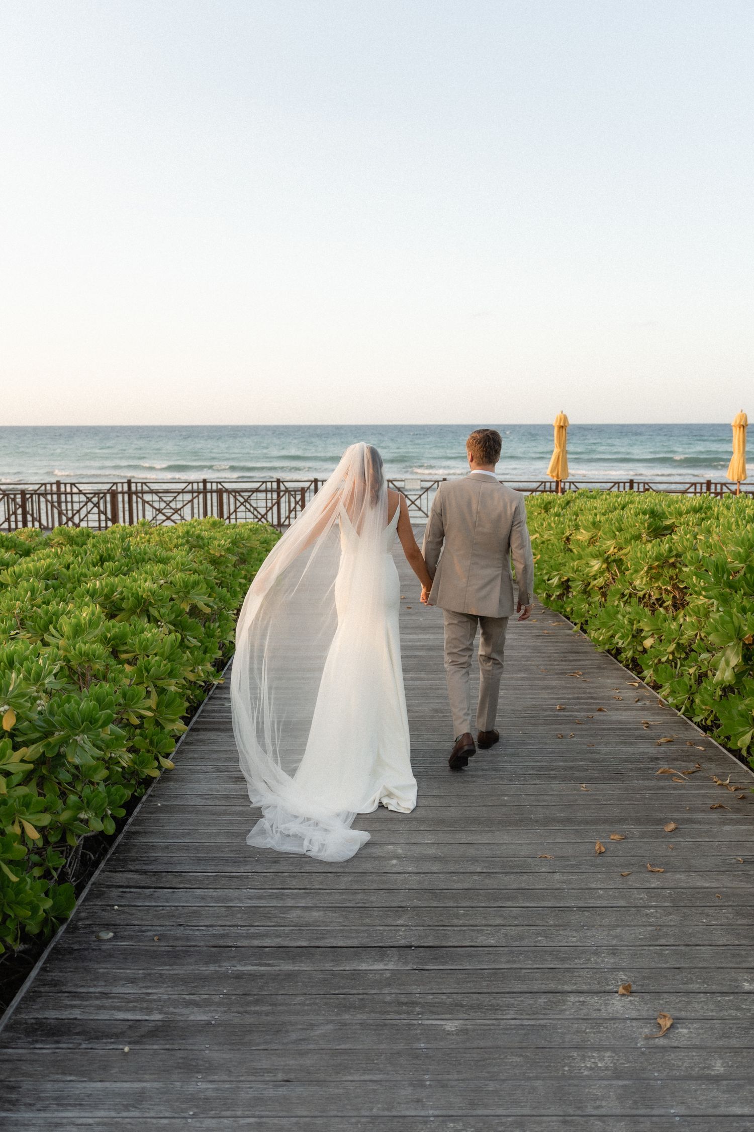 Couple walking along a wooden boardwalk by the ocean during sunset at a tropical resort.