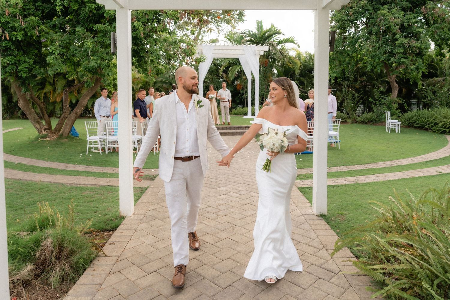 A wedding couple walks along a white columned pathway in a garden setting.