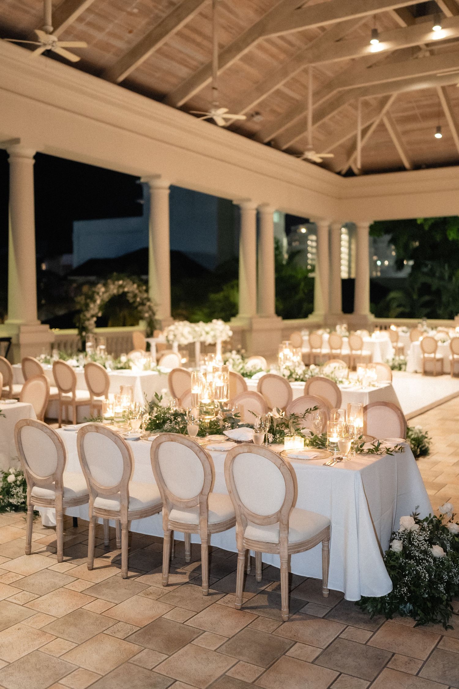 Elegant wedding reception setup with white chairs and tables under a covered outdoor pavilion.