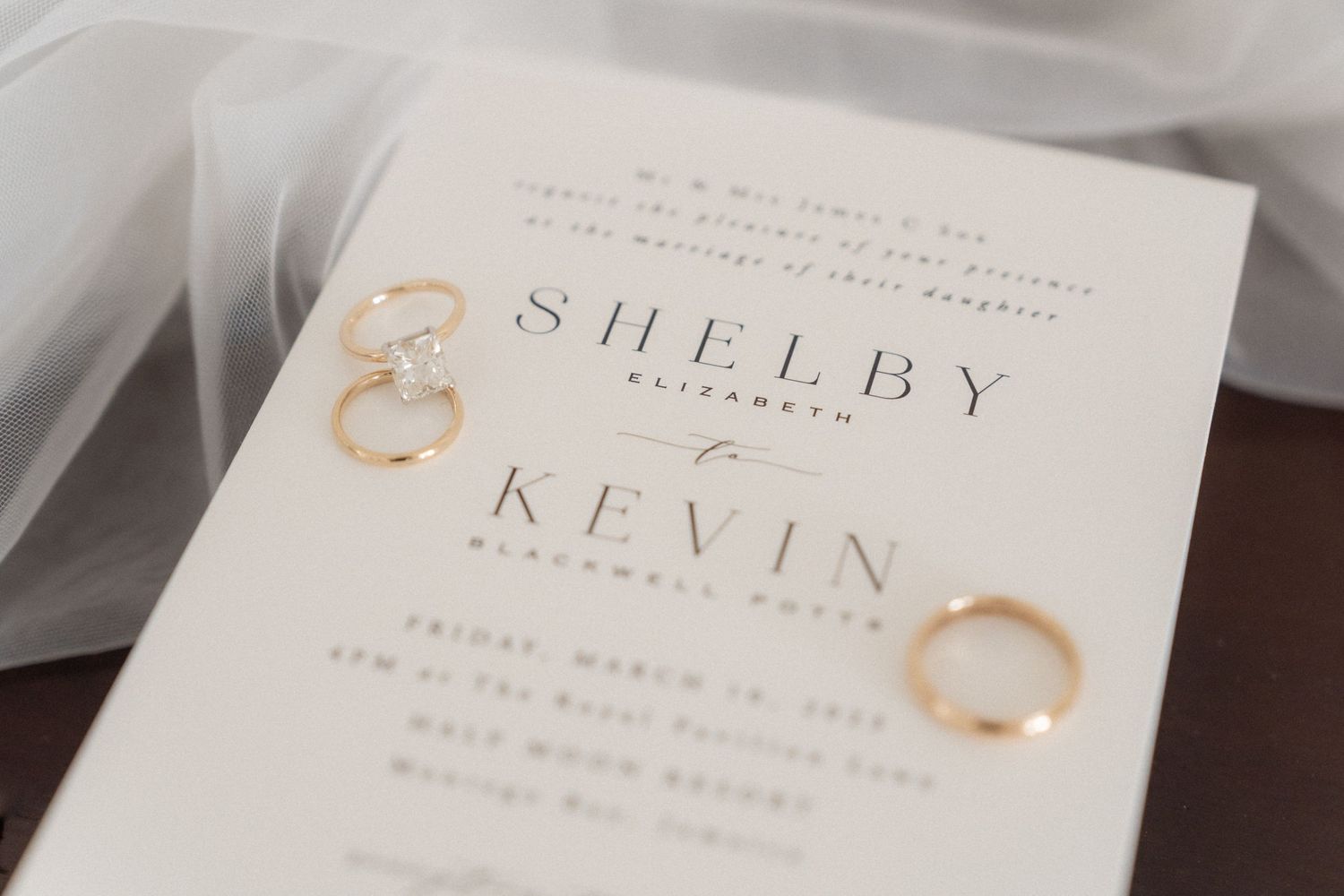 Wedding invitation with gold rings showing names Shelby and Kevin on elegant white paper.