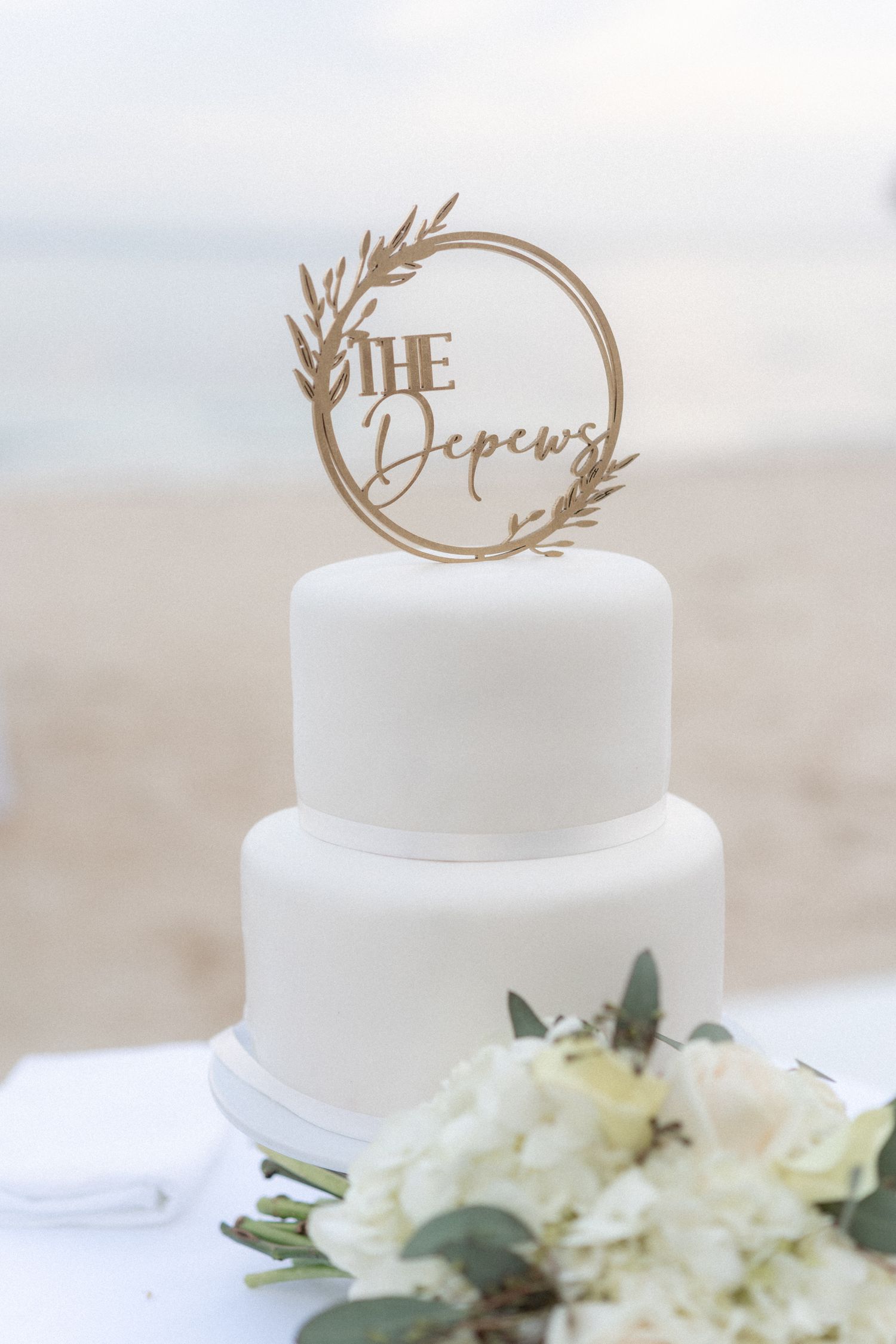 Simple two-tier white wedding cake with custom wooden name topper and white floral decoration.