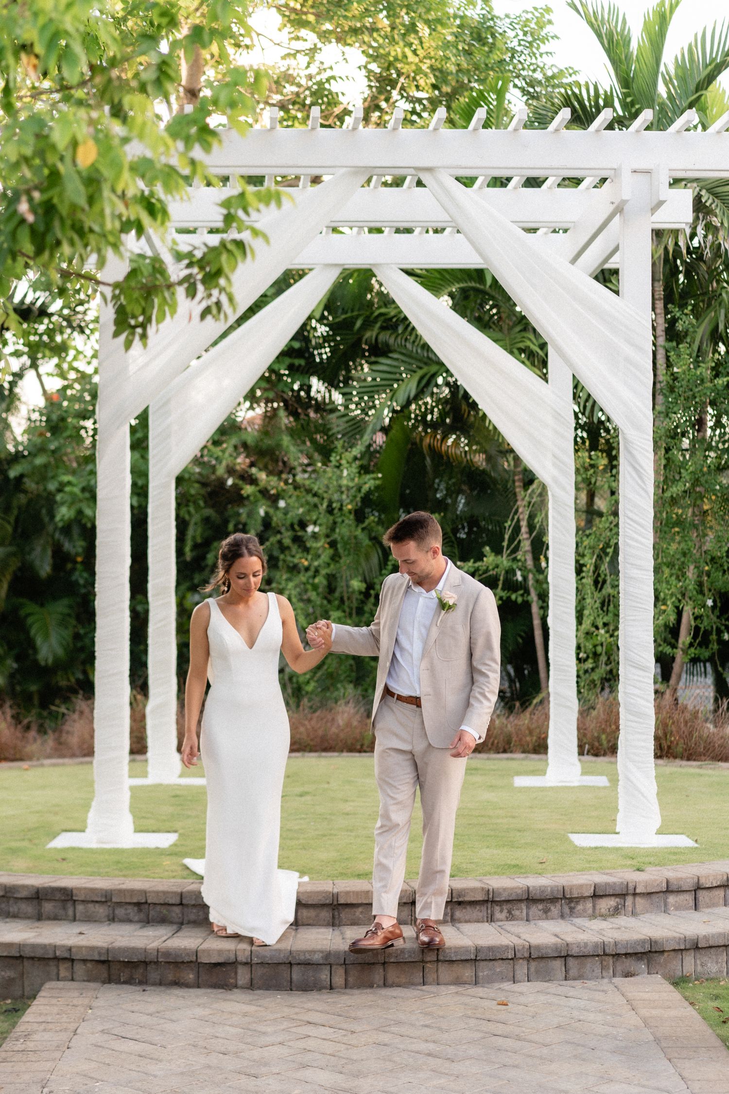 Couple stands under white wooden wedding arch on elevated stone platform.