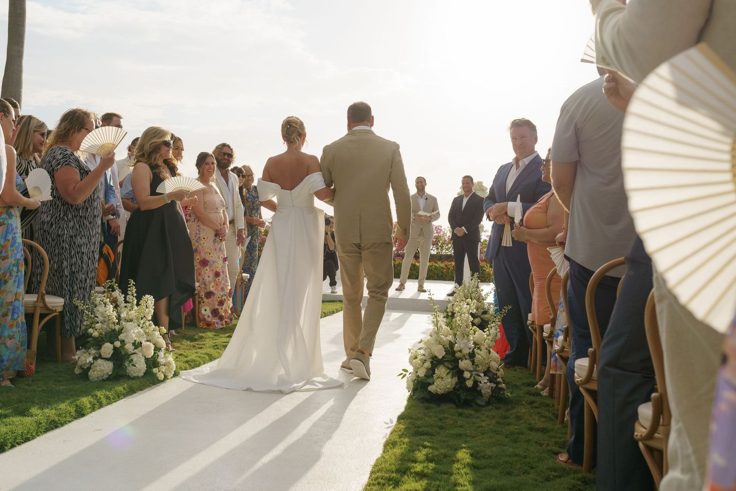 A couple walks down an outdoor wedding aisle lined with guests and floral arrangements during a sunny ceremony.