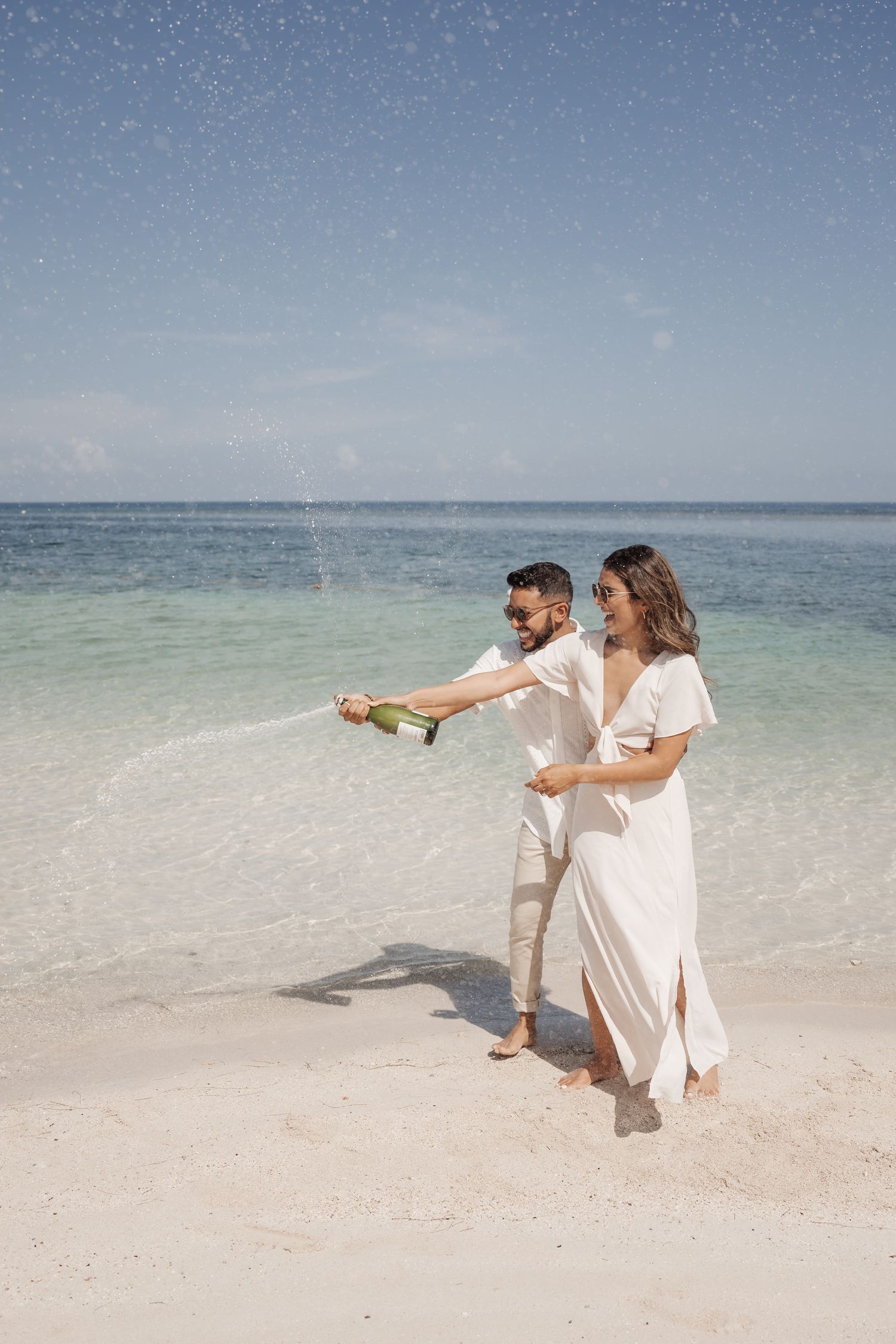 A couple dances playfully together on a white sandy beach with turquoise waters in the background.