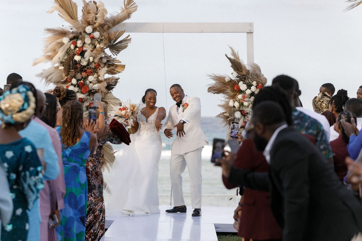 Wedding celebration with guests cheering under an elegant white arch with dried flowers.