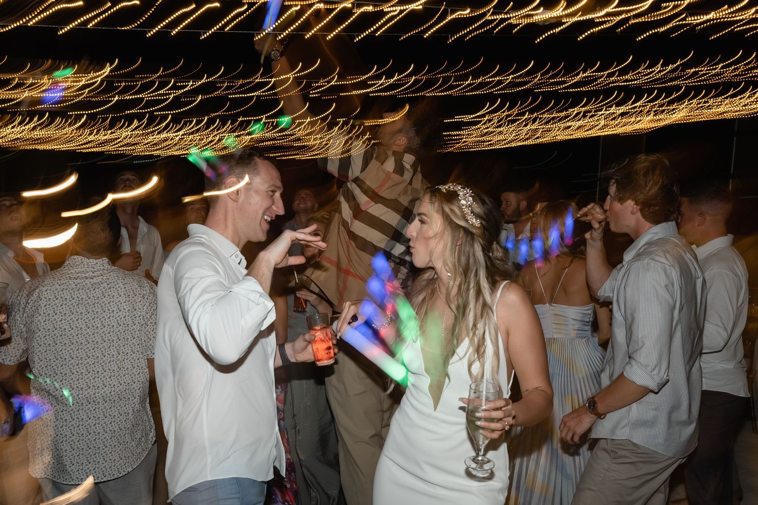 Wedding reception guests dancing and holding colorful light-up sticks during evening celebrations.