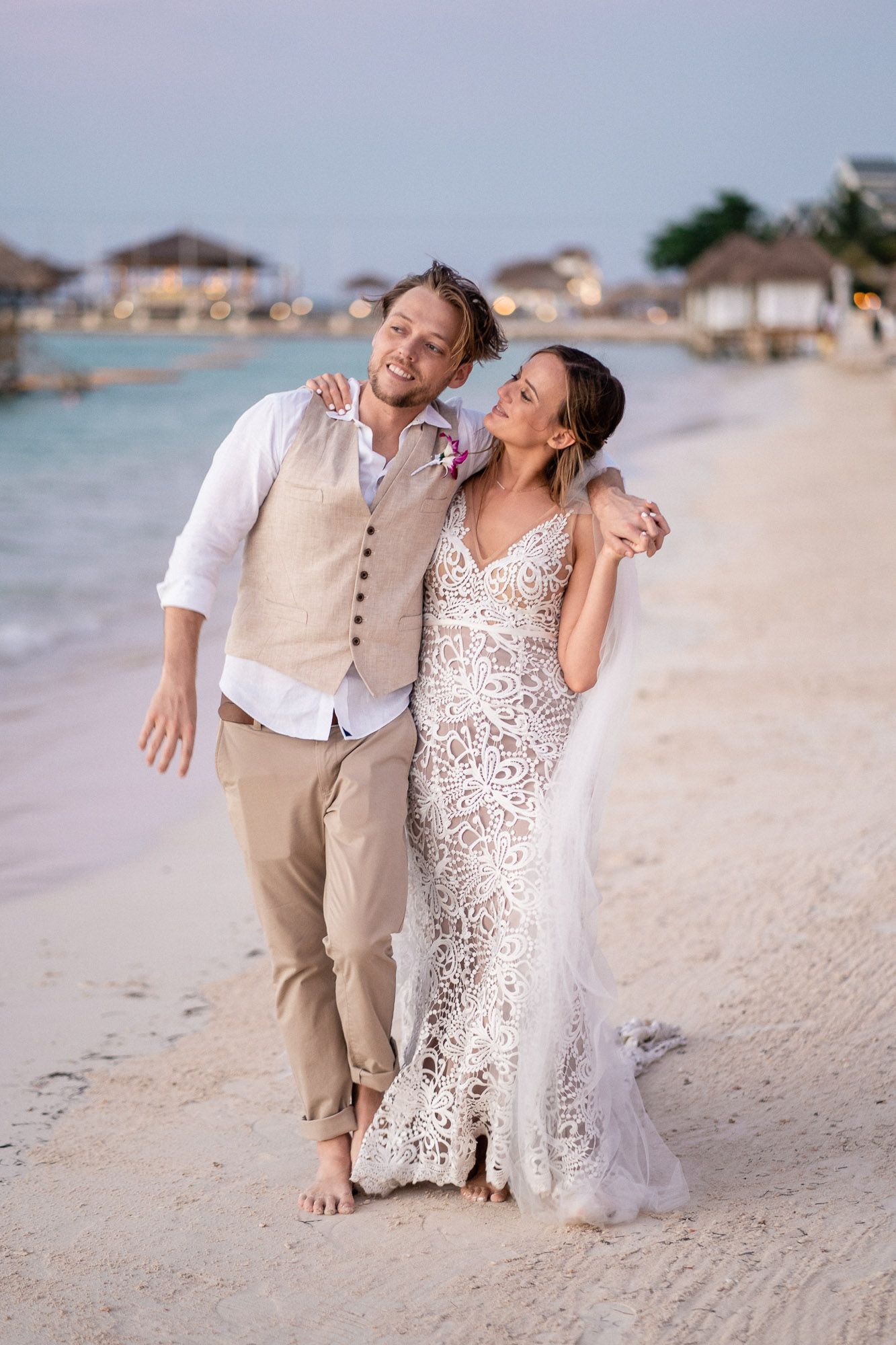 A joyful beach wedding couple strolls along a tropical shoreline with resort bungalows in the background.
