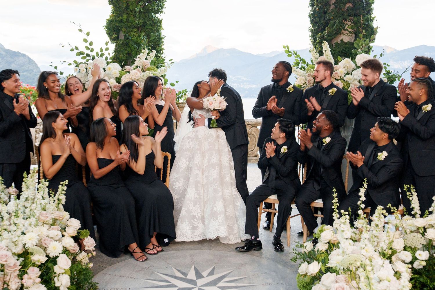 Wedding guests in black attire gather around a floral-decorated outdoor venue with mountain views in the background.
