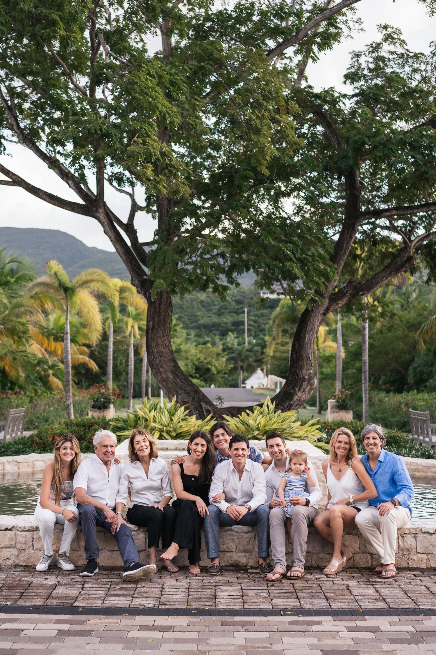 Extended family gathering outdoors on stone wall with tropical trees and mountains in background.